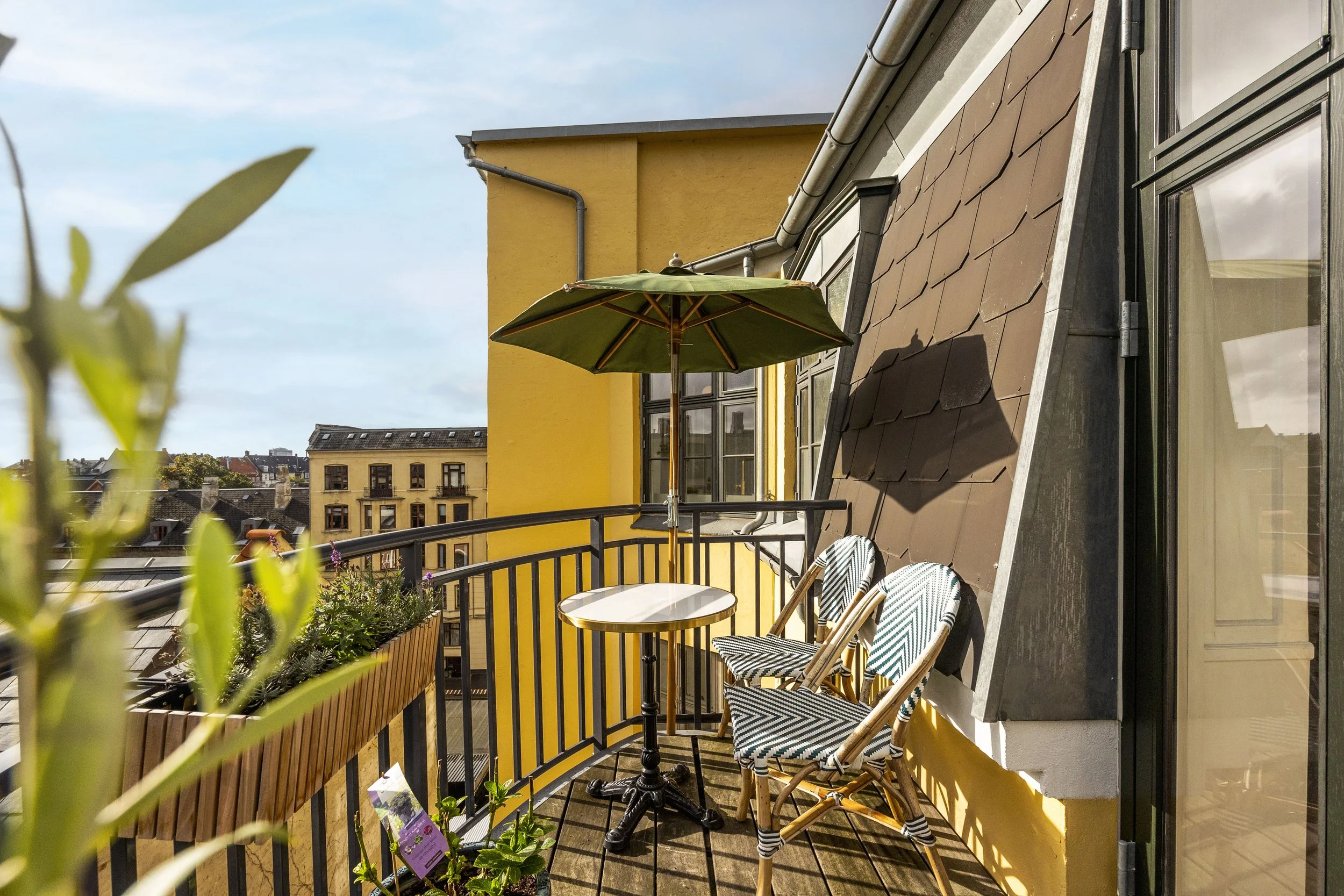Balcony with yellow exterior wall, table, two striped chairs, green umbrella, outdoor plants, city buildings in background.
