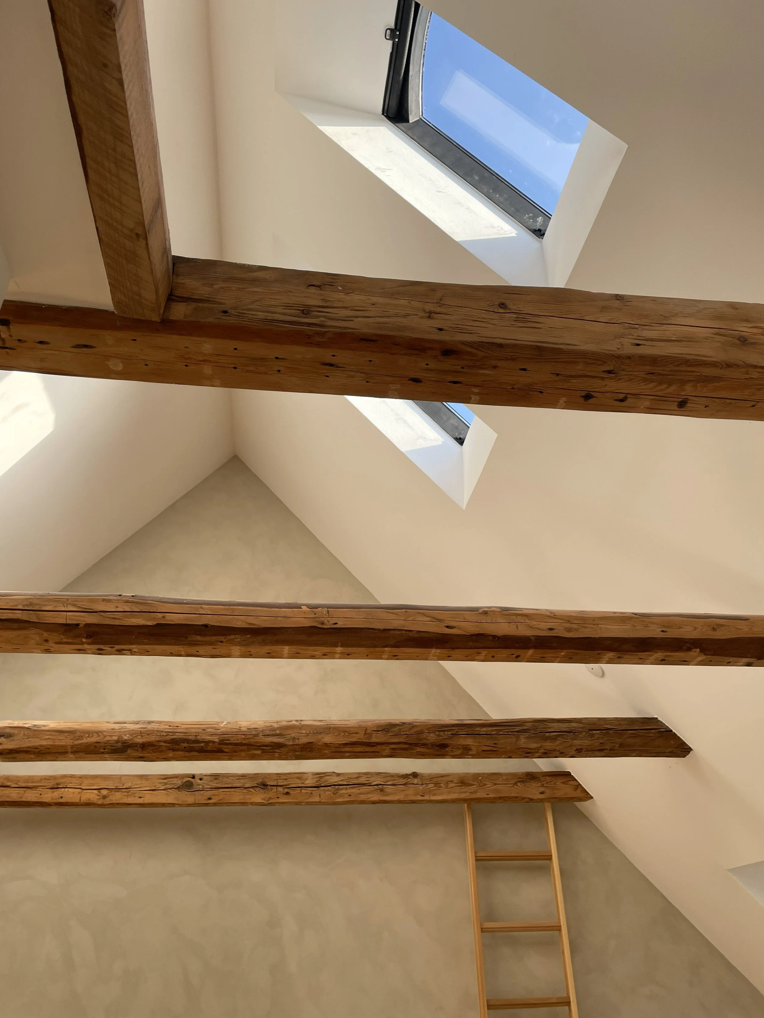View of a room with sloped ceilings, exposed wooden beams, and three skylight windows showing a blue sky.