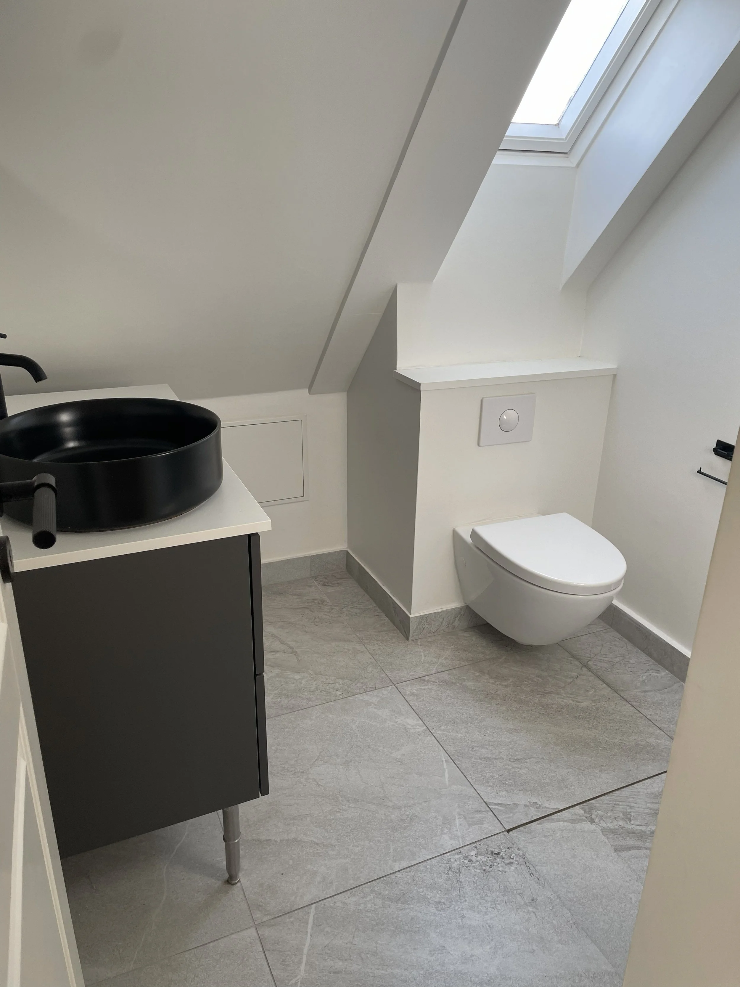 Small modern bathroom with white walls and sloped ceiling, featuring a wall-mounted toilet, a black sink on a white vanity, and a skylight window letting in natural light.