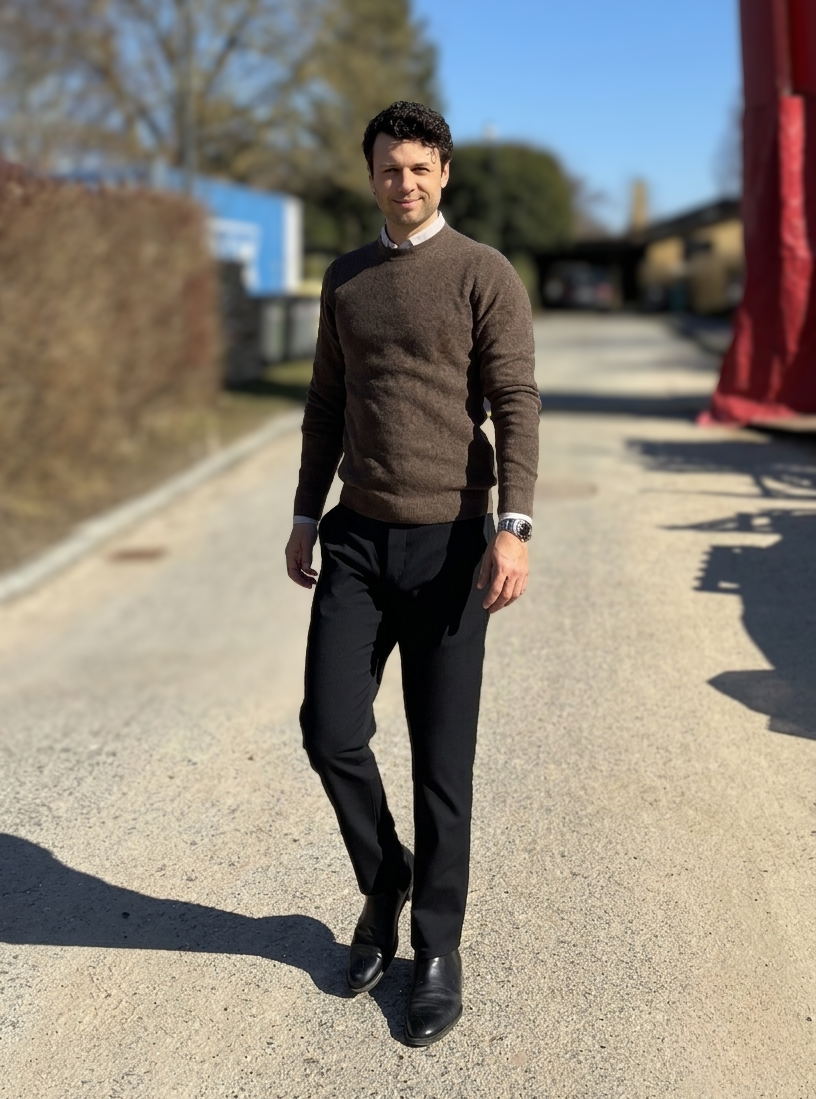 A man walking outdoors on a sunny day, wearing a brown sweater, black pants, and black shoes, with hills and trees in the background.