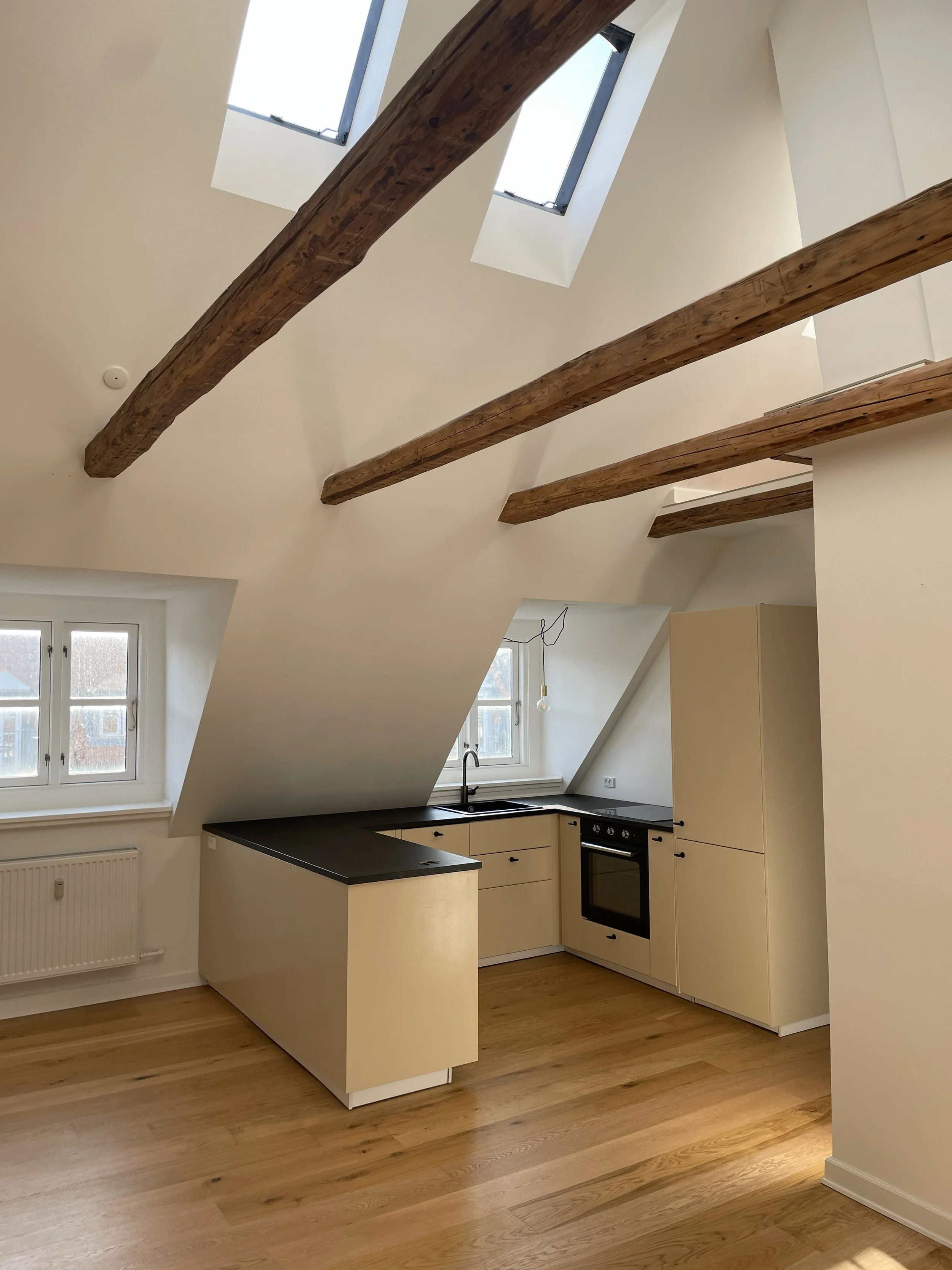 Empty attic kitchen with sloped ceiling, exposed wooden beams, two skylights, a window, light beige cabinets, black countertops, a black oven, a sink, and wood flooring.