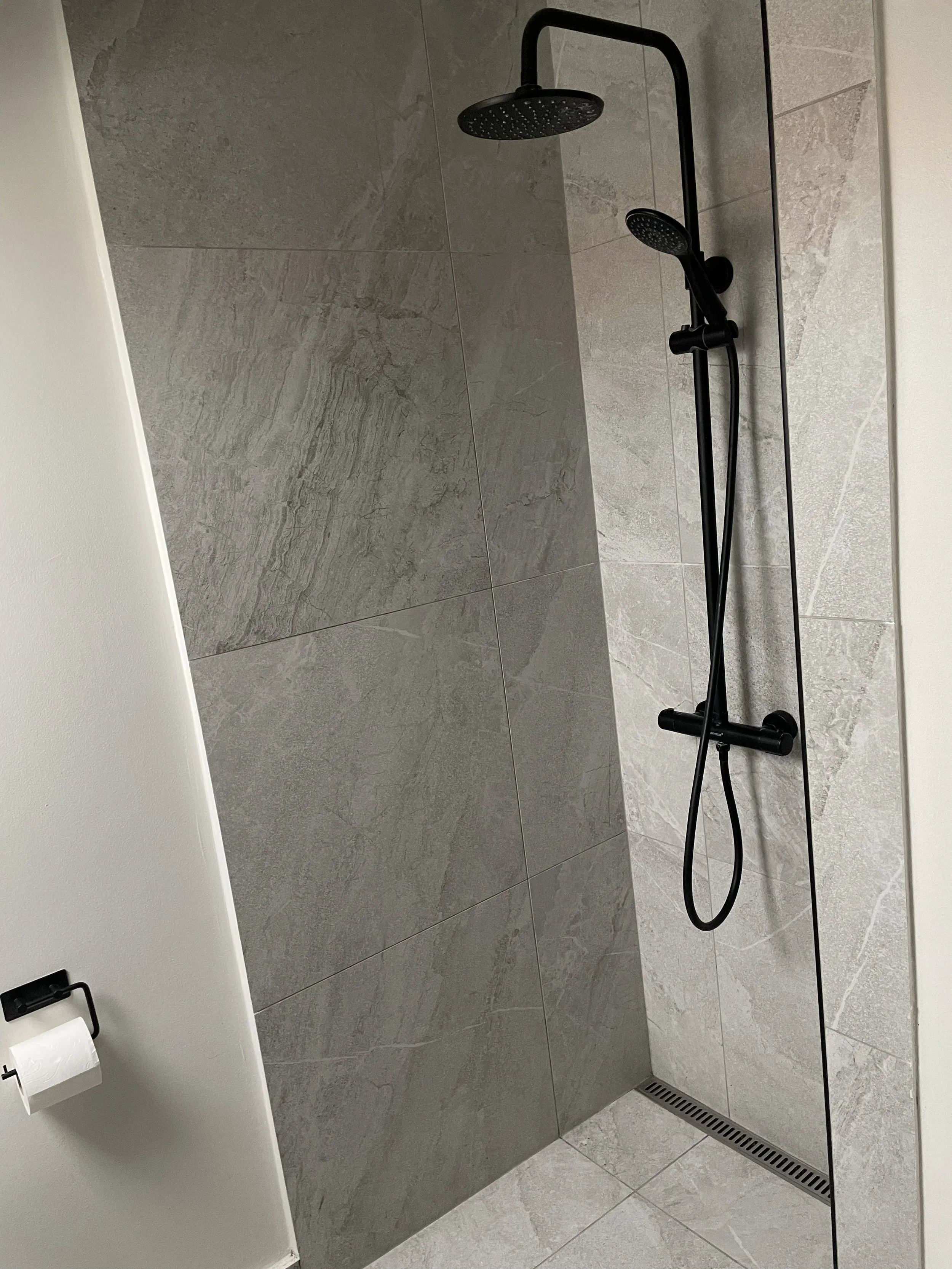 A modern shower area with large gray tiles, a black rainfall showerhead, a handheld shower wand, and a linear floor drain.