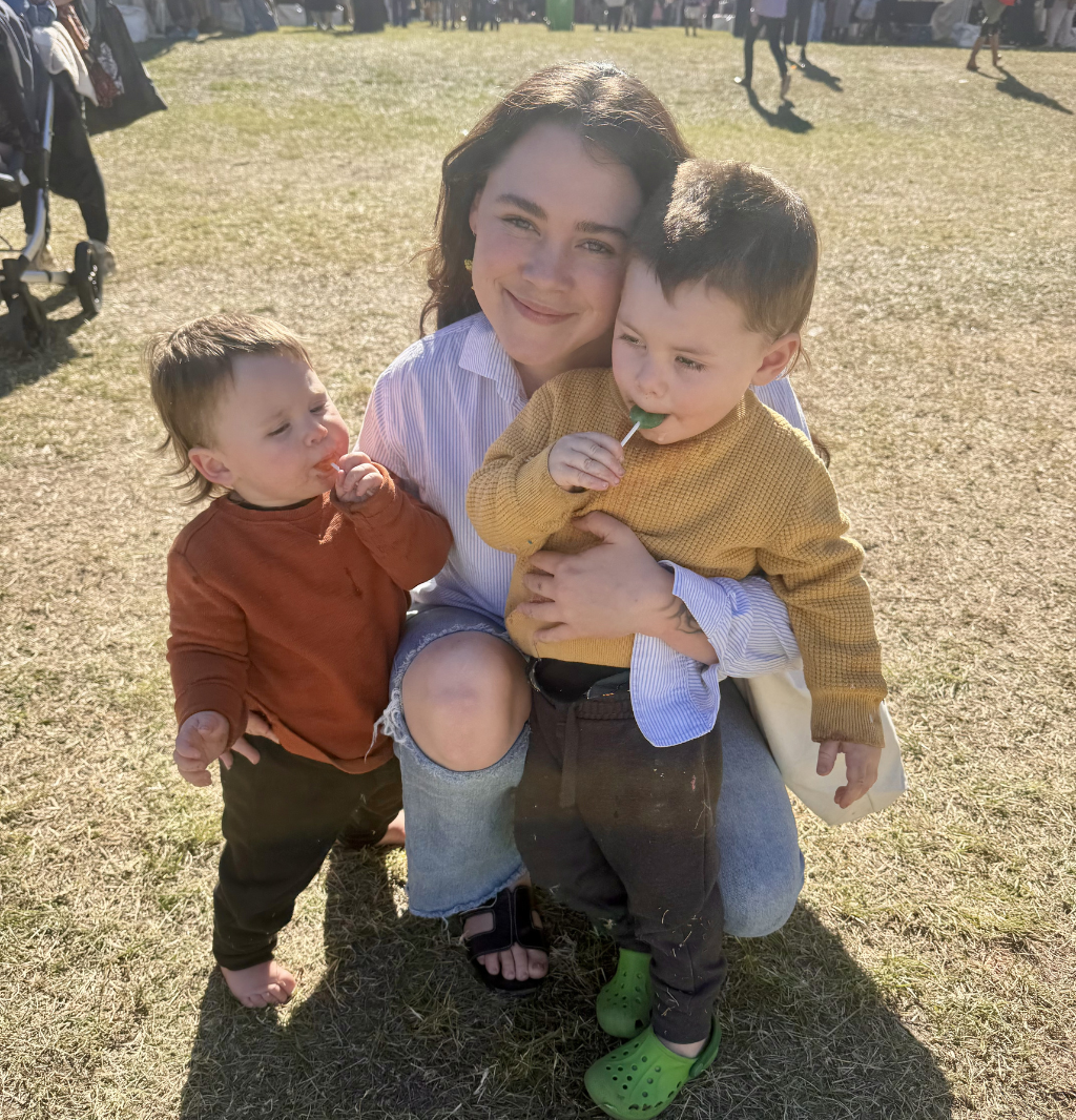 A young woman with dark wavy hair smiling, holding two children in an outdoor setting on a sunny day. One child has short dark hair and is wearing a mustard yellow sweater, eating a green lollipop. The other child has light brown hair, wearing a reddish-brown top and black pants, and is also holding a lollipop.