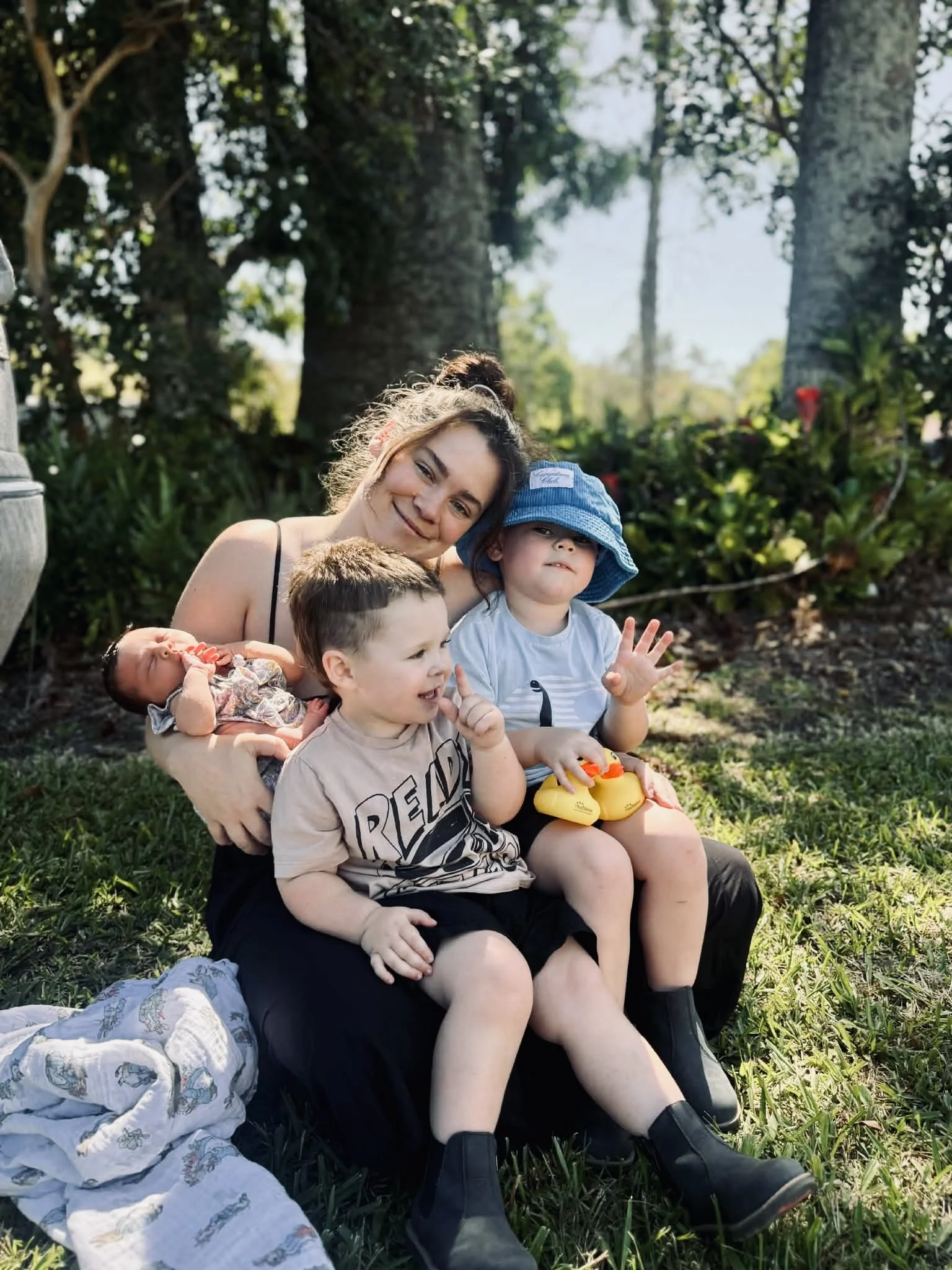 Young woman with brown hair in a bun sitting on grass in a park with three small children, two boys and a girl, all smiling and sitting close together. The girl is wearing a blue sun hat, one boy has a beige T-shirt with text, and the other boy is holding a stuffed animal. Background features trees and greenery.