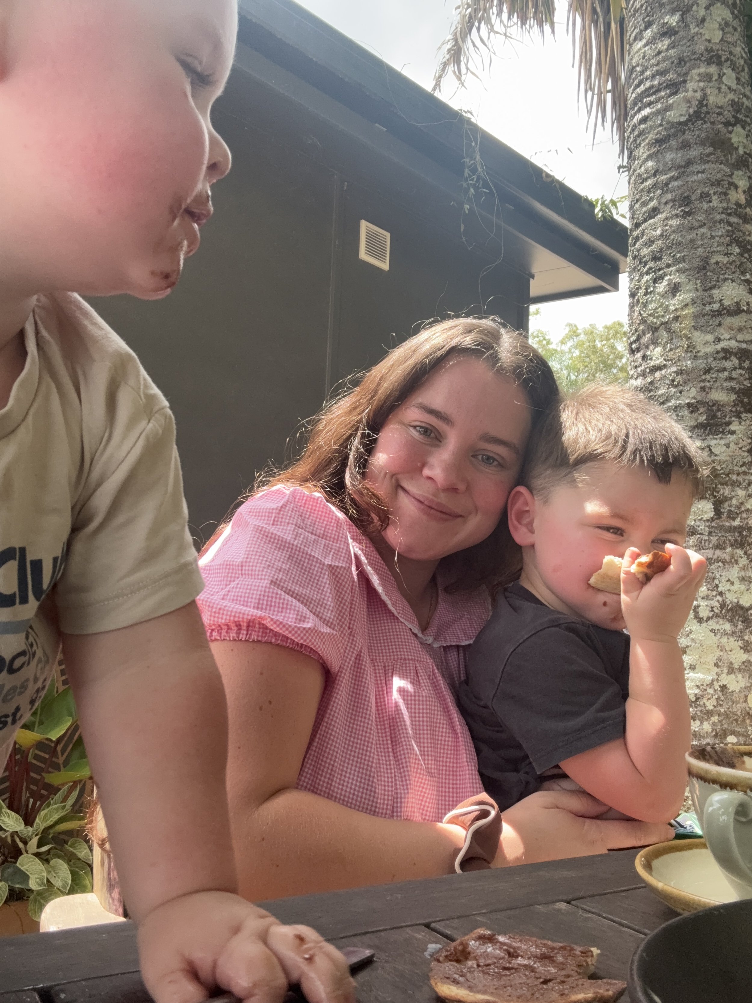 A woman with brown hair, wearing a pink checkered shirt, is smiling at the camera as she sits outdoors with two children. One child, with light hair, is in the foreground and partially visible, and the other, a young boy with short brown hair, is holding a slice of pizza. They are sitting at a table with cups and a piece of toast. There is a palm tree and a dark building in the background.