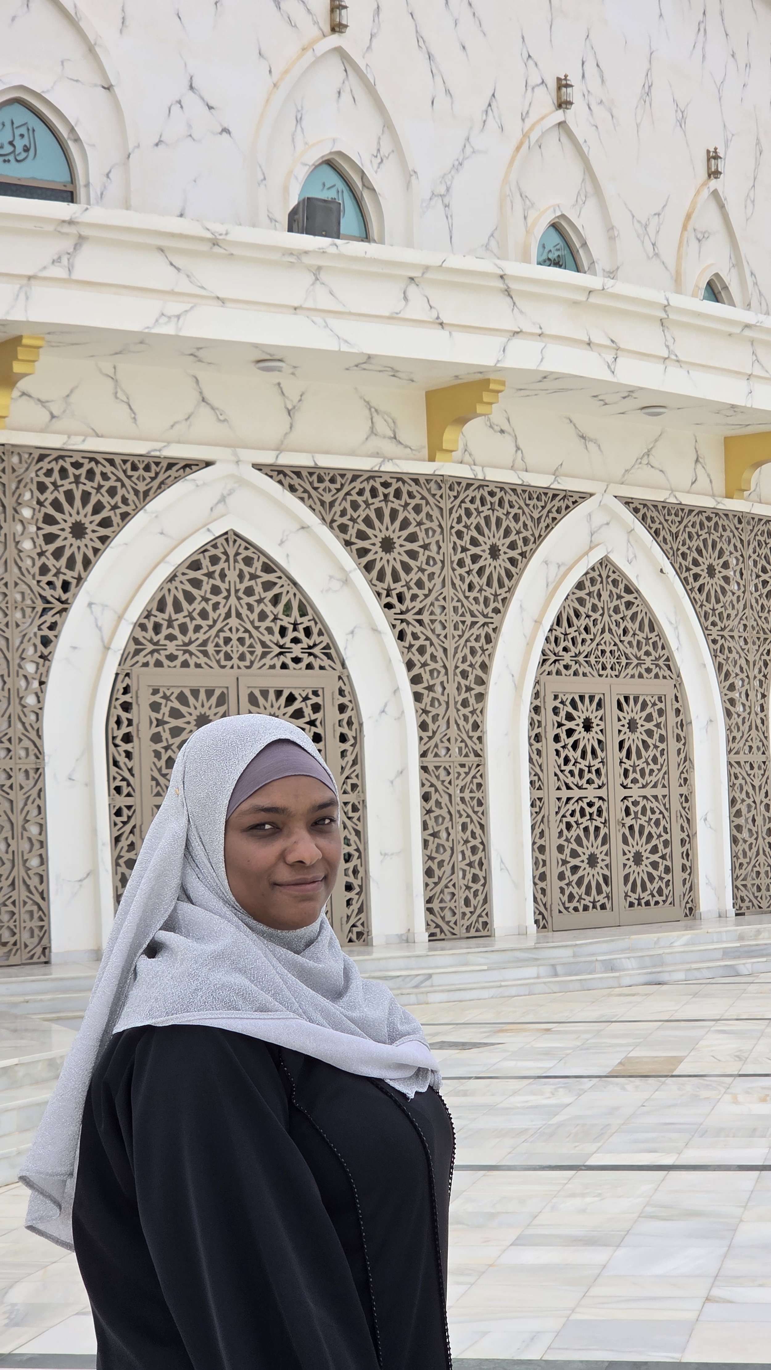 A woman wearing a headscarf standing in front of a white marble building with intricate Islamic architectural elements.