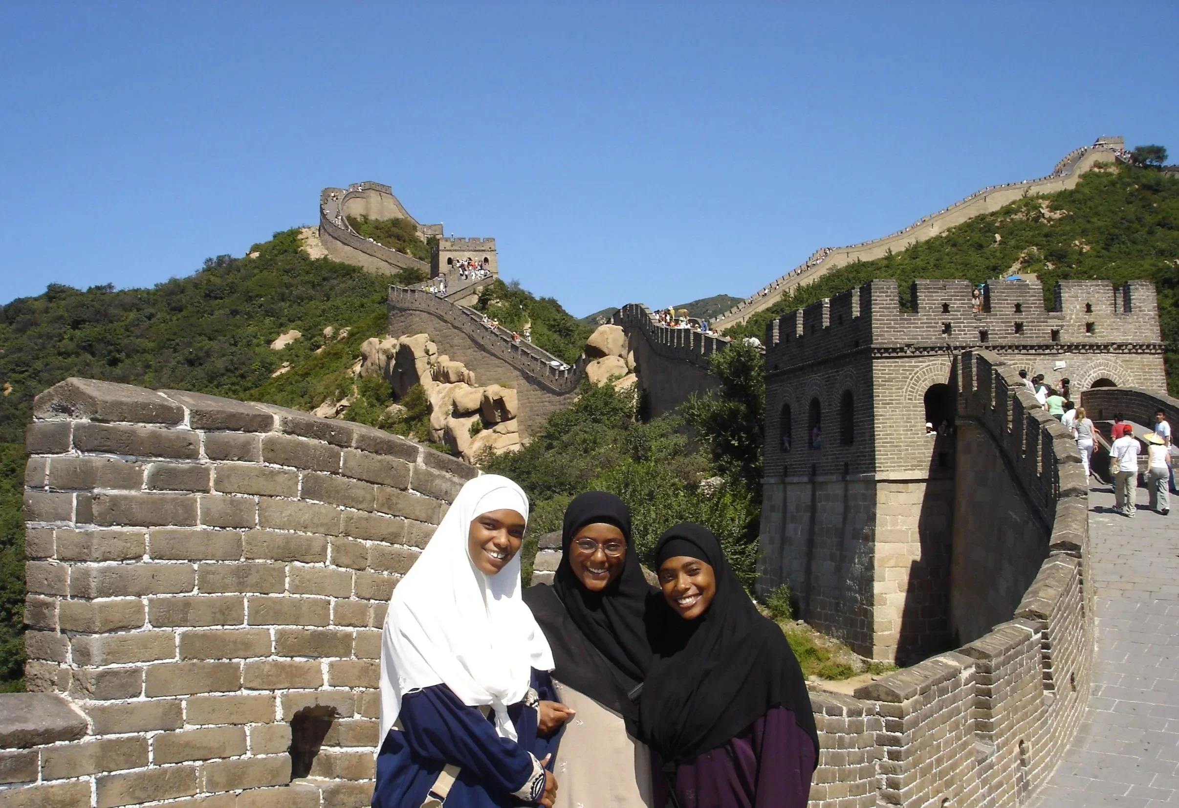 Three women in hijabs and modest clothing smiling at the camera on the Great Wall of China with clear sky and green hills in the background.
