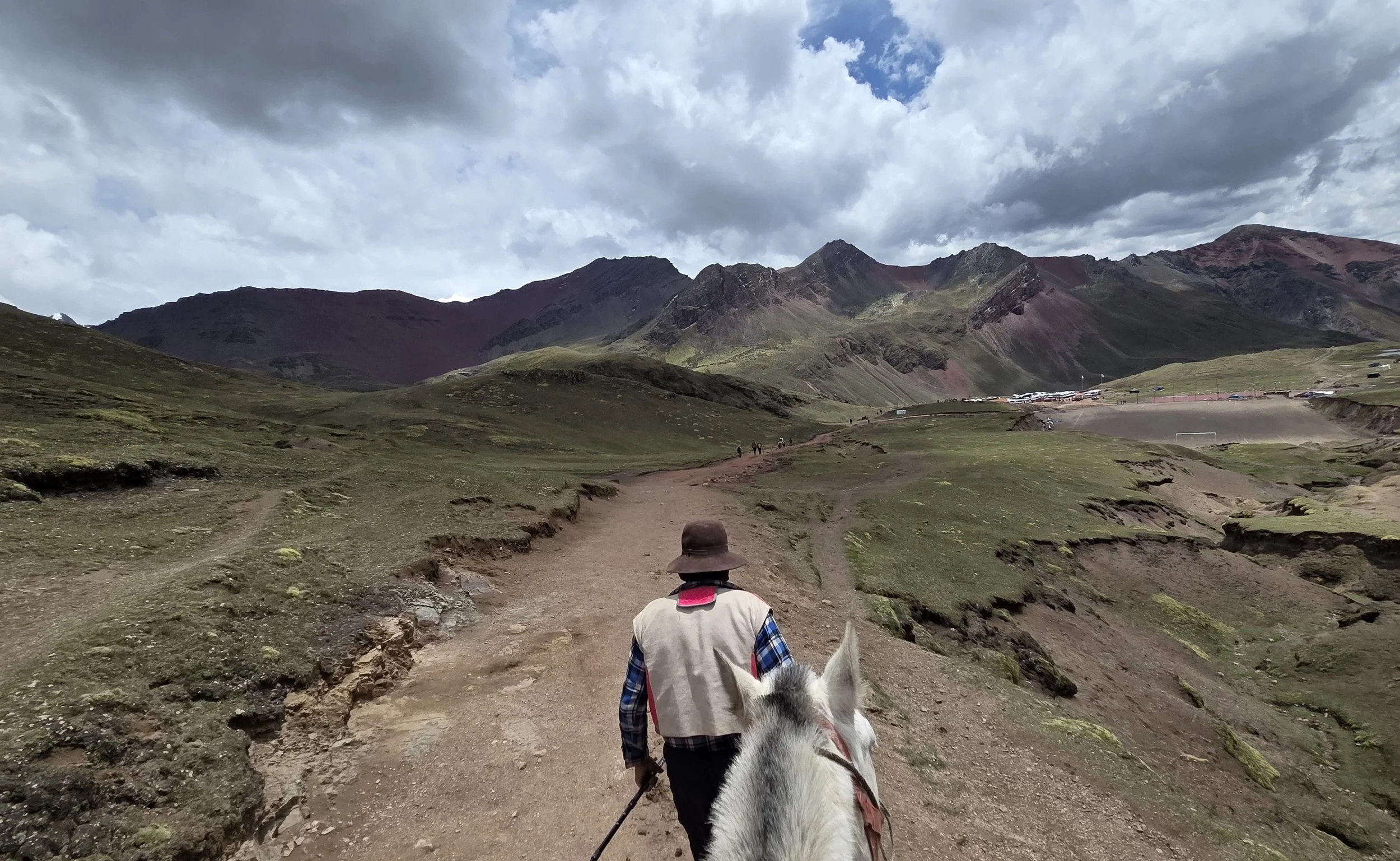 A person riding a white horse along a dirt trail in a mountainous landscape. The rider is wearing a wide-brimmed hat, a beige vest, and a plaid shirt, with mountains and cloudy sky in the background.