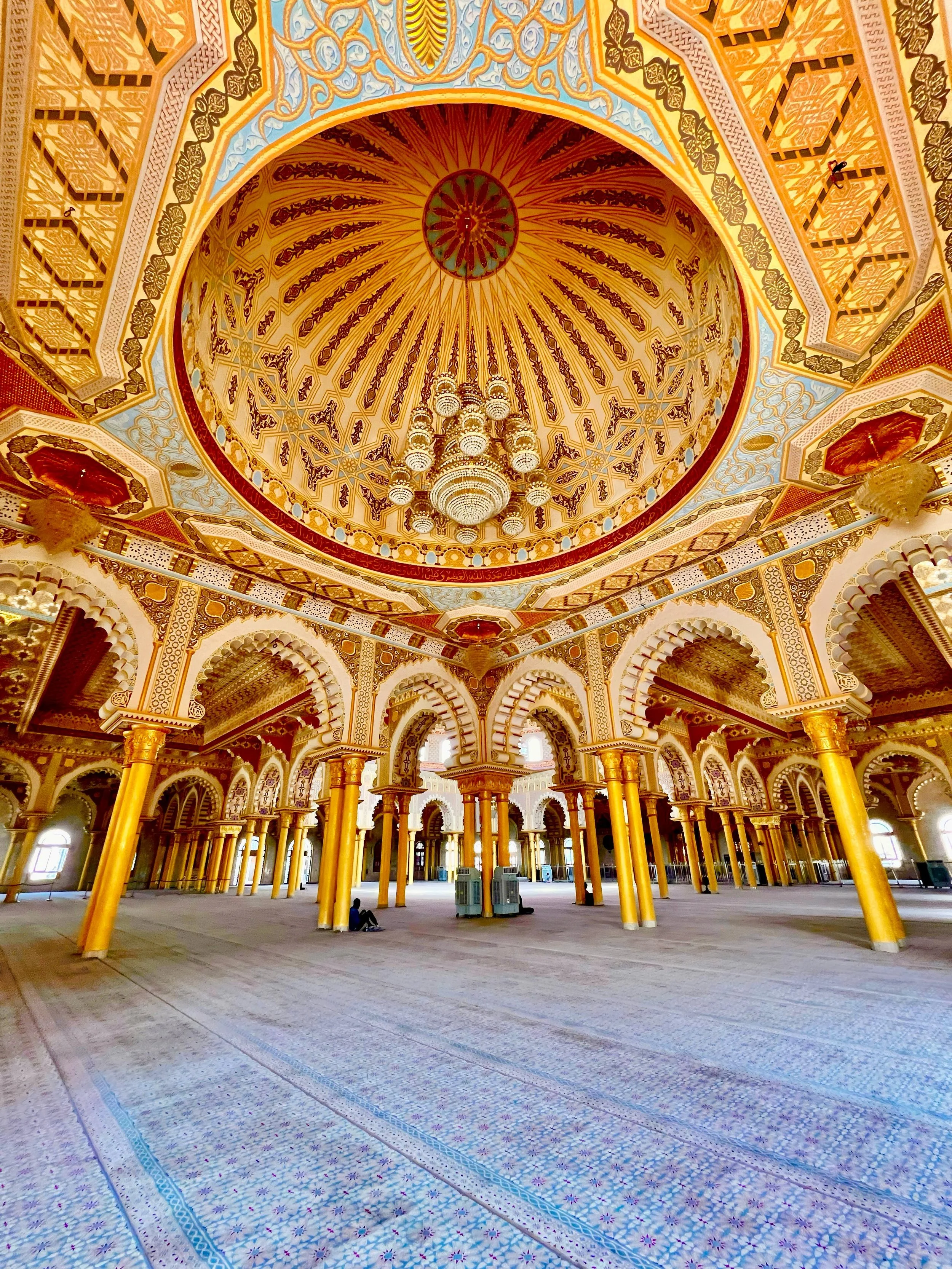Interior of a grand mosque with ornate gold and red decorations, Arabic-style arches, a large chandelier hanging from a decorated domed ceiling, and patterned carpeted floors.