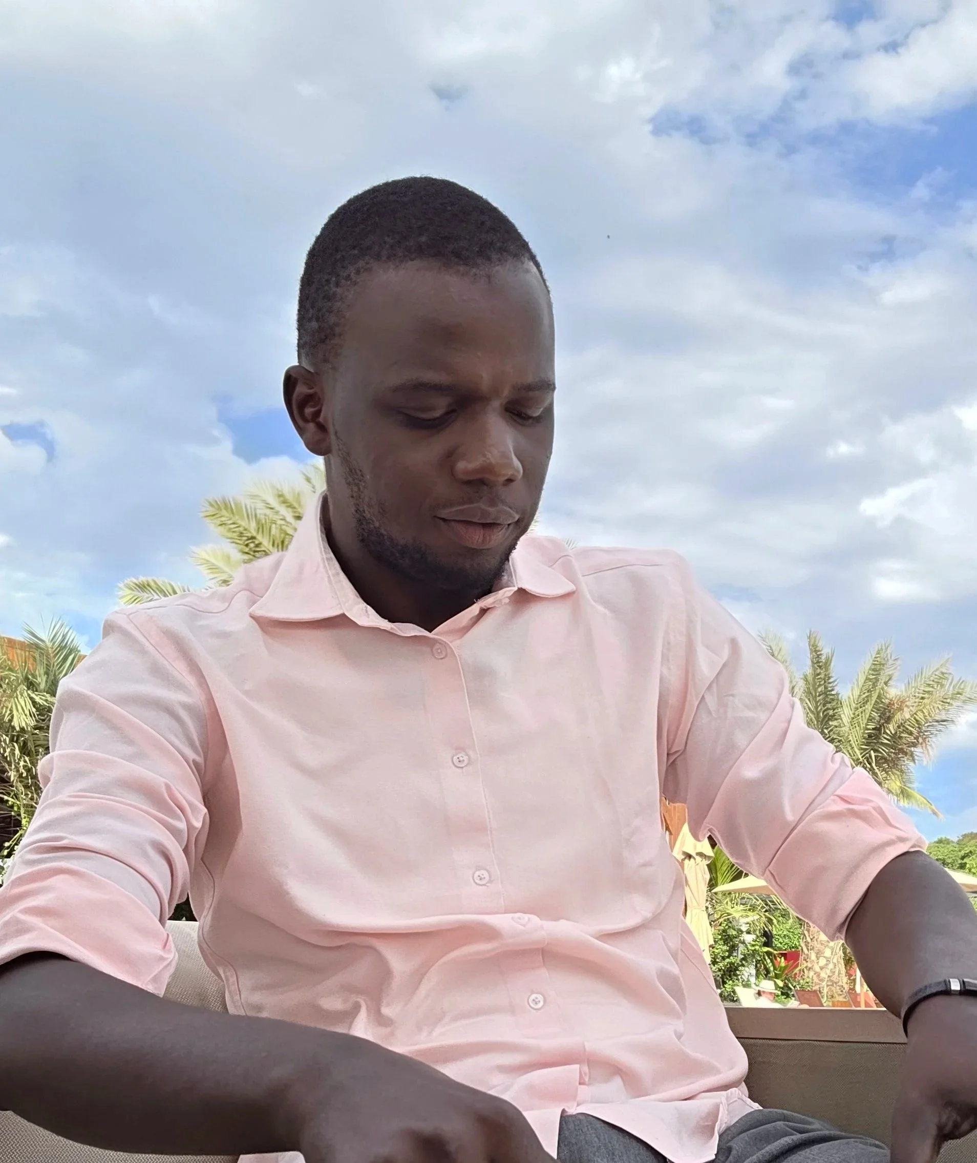 A man in a light pink button-up shirt sitting outdoors with a partly cloudy sky and palm trees in the background.