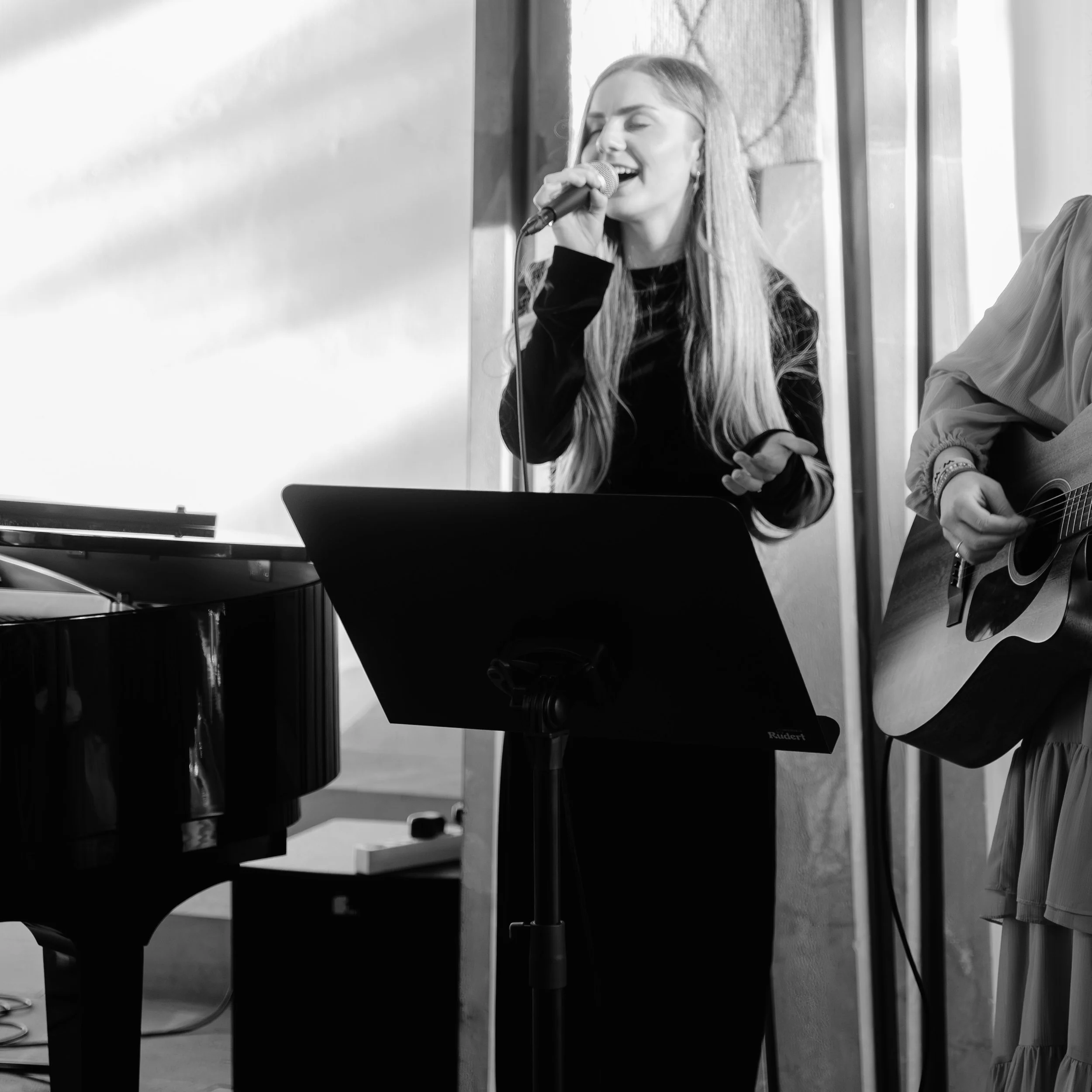 Eine junge Frau singt mit einem Mikrofon, während eine andere Person Gitarre spielt, in einem Raum mit großem Fenster im Hintergrund.