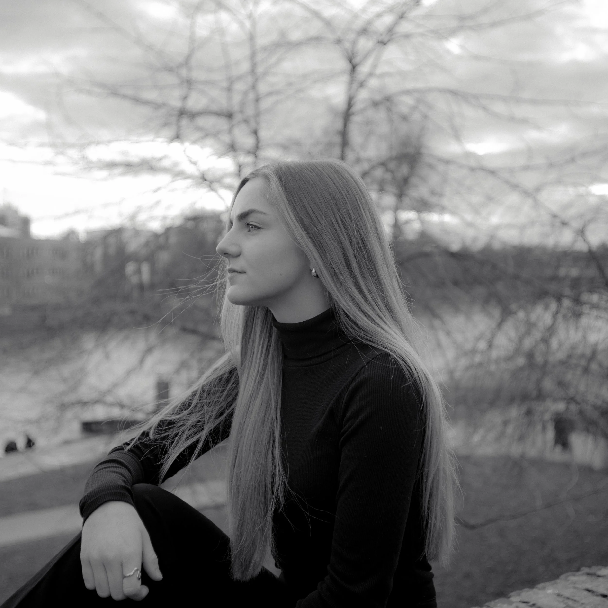 Schwarz-Weiß-Fotografie einer jungen Frau in Profilansicht bei Sonnenuntergang, sitzend mit Blick nach links, im Hintergrund Bäume und Gebäude.