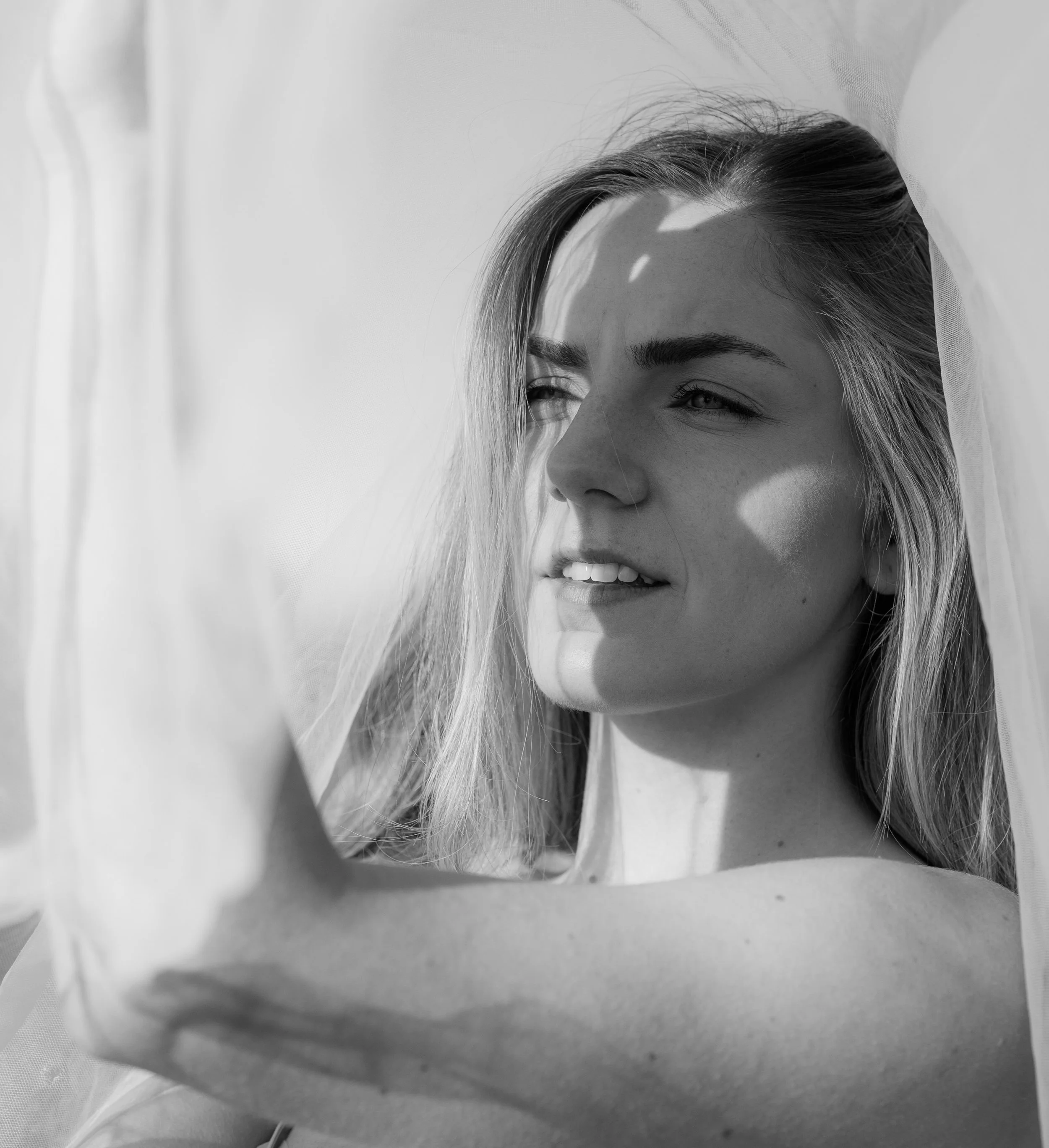 Schwarz-weiß Porträt einer jungen Frau mit langem Haar, Sonnenschein auf ihrem Gesicht, halb verdeckt durch einen Schleier oder Tüll, Blick nach rechts, entspannte Stimmung.