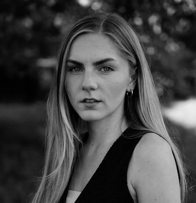 Schwarz-weiß-Bild eines jungen Mädchens mit langen, glatten Haaren, trägt ein kleines Hufeisen-Ohrring und ein ärmelloses Oberteil, blickt ernst in die Kamera vor unscharfem Hintergrund.