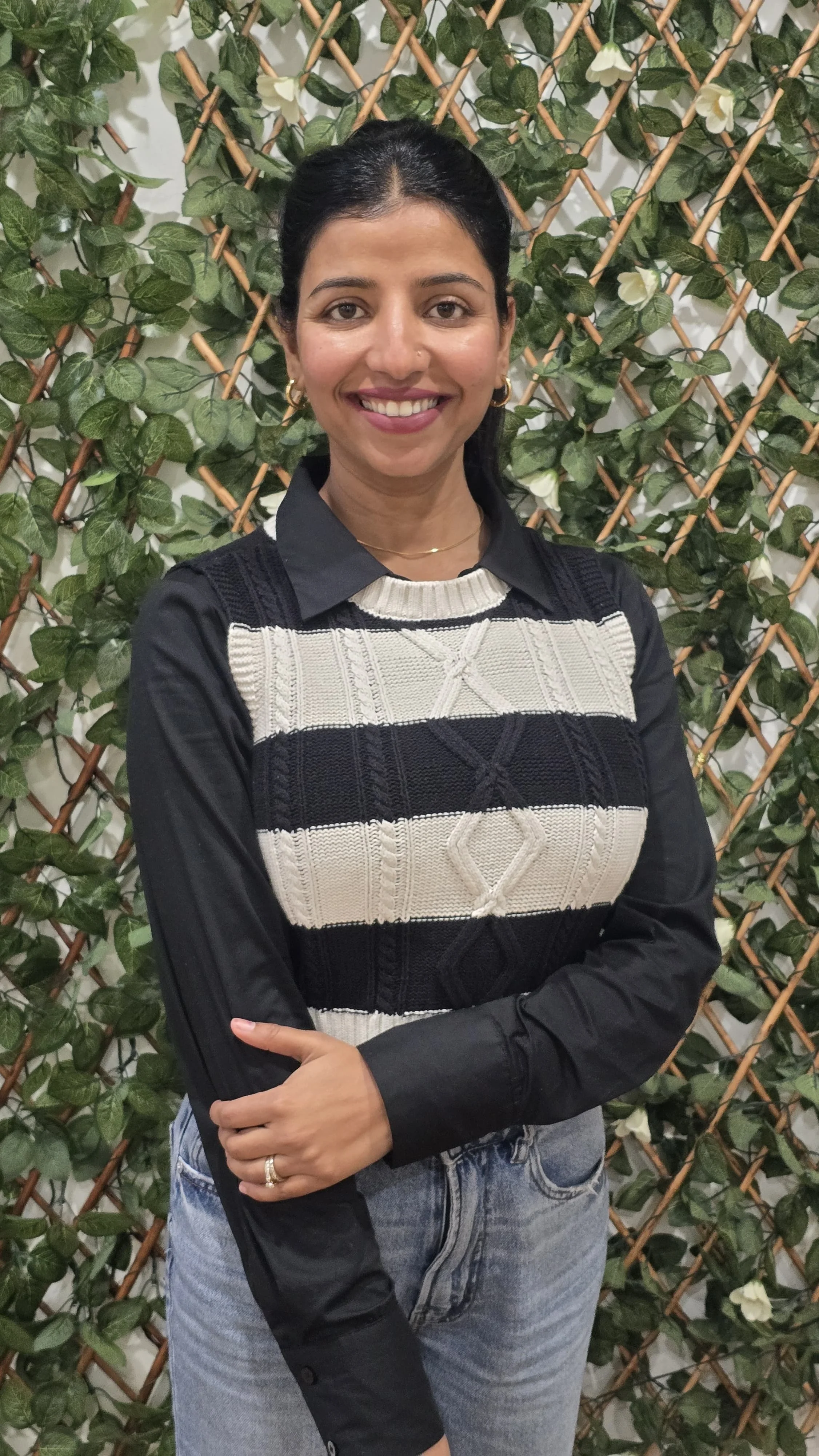 A woman with dark hair pulled back, wearing a black and white patterned sweater over a black shirt, and light blue jeans, stands in front of a leafy green wall with wooden lattice.