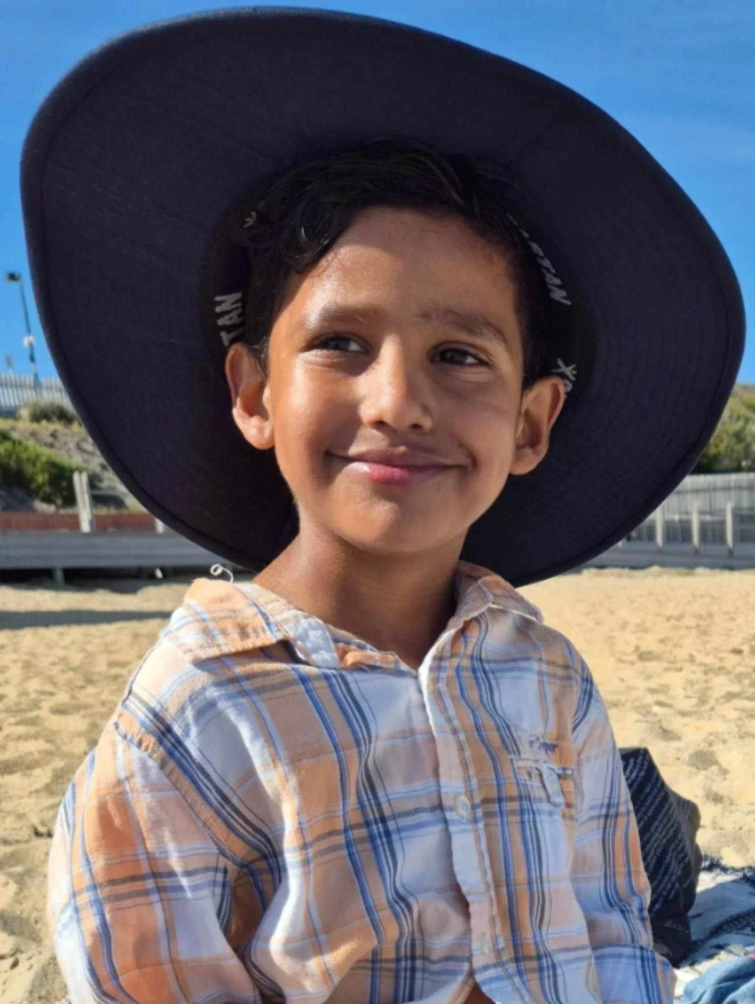 Close-up photo of a smiling boy wearing a large black hat with a wide brim, outdoors on a sunny day, with a fence and blue sky in the background.