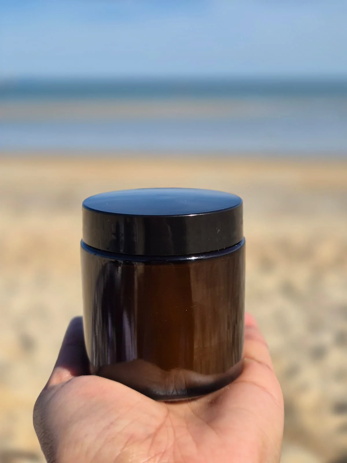 A hand holding a brown insulated tumbler with a black lid on a sandy beach with the ocean and blue sky in the background.