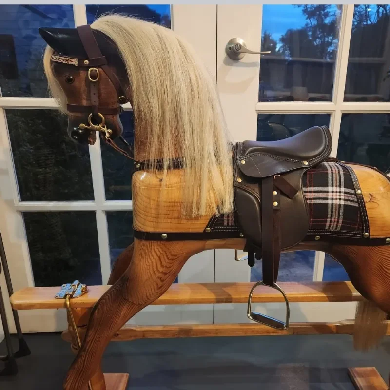 A wooden rocking horse with a blonde horse's mane, equipped with a black saddle and plaid saddle blanket, placed on a wooden bench in front of a glass door with a window showing trees outside.
