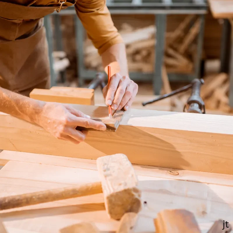 Close-up of a person marking a wooden plank with a pencil in a woodworking shop, with tools and wood in the background.