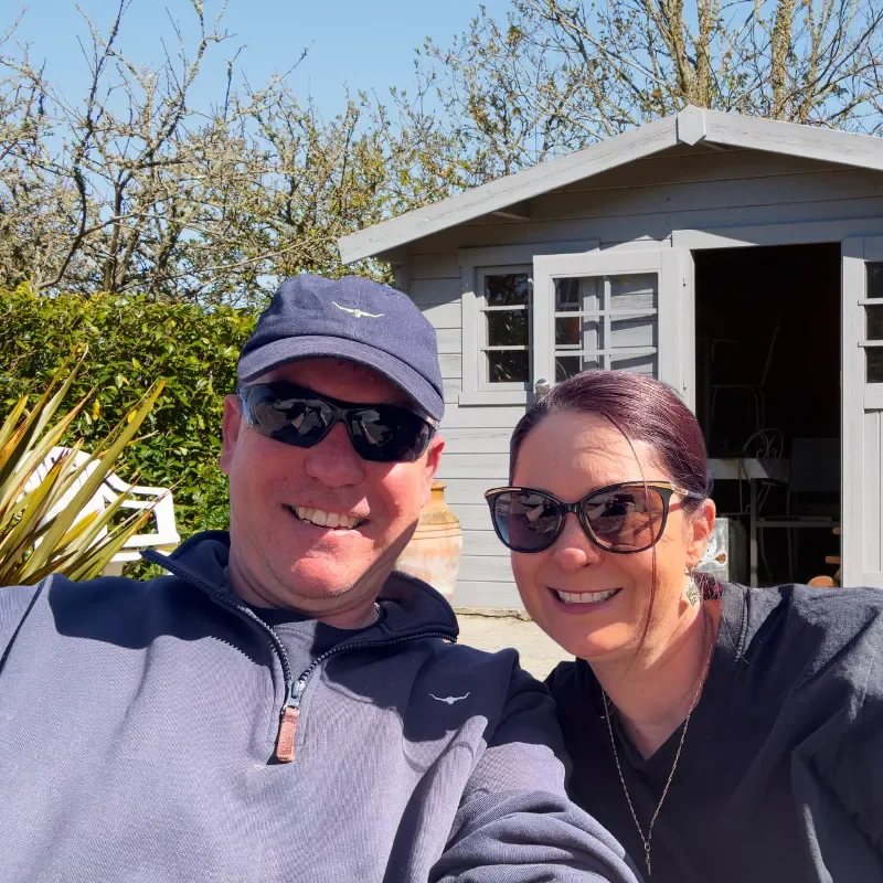 A smiling man and woman wearing sunglasses outdoors in front of a small gray shed, with trees and blue sky in the background.