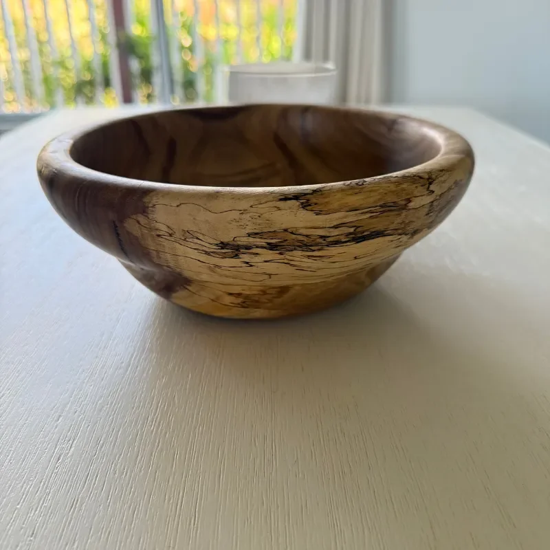 A wooden bowl with visible grain patterns sitting on a white surface, with a blurred background showing a window and outdoor scenery.