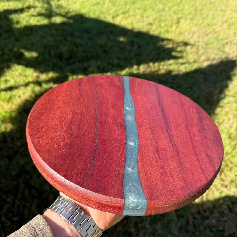 Hand holding a round wooden table with a blue resin river in the middle, outdoors on grass.