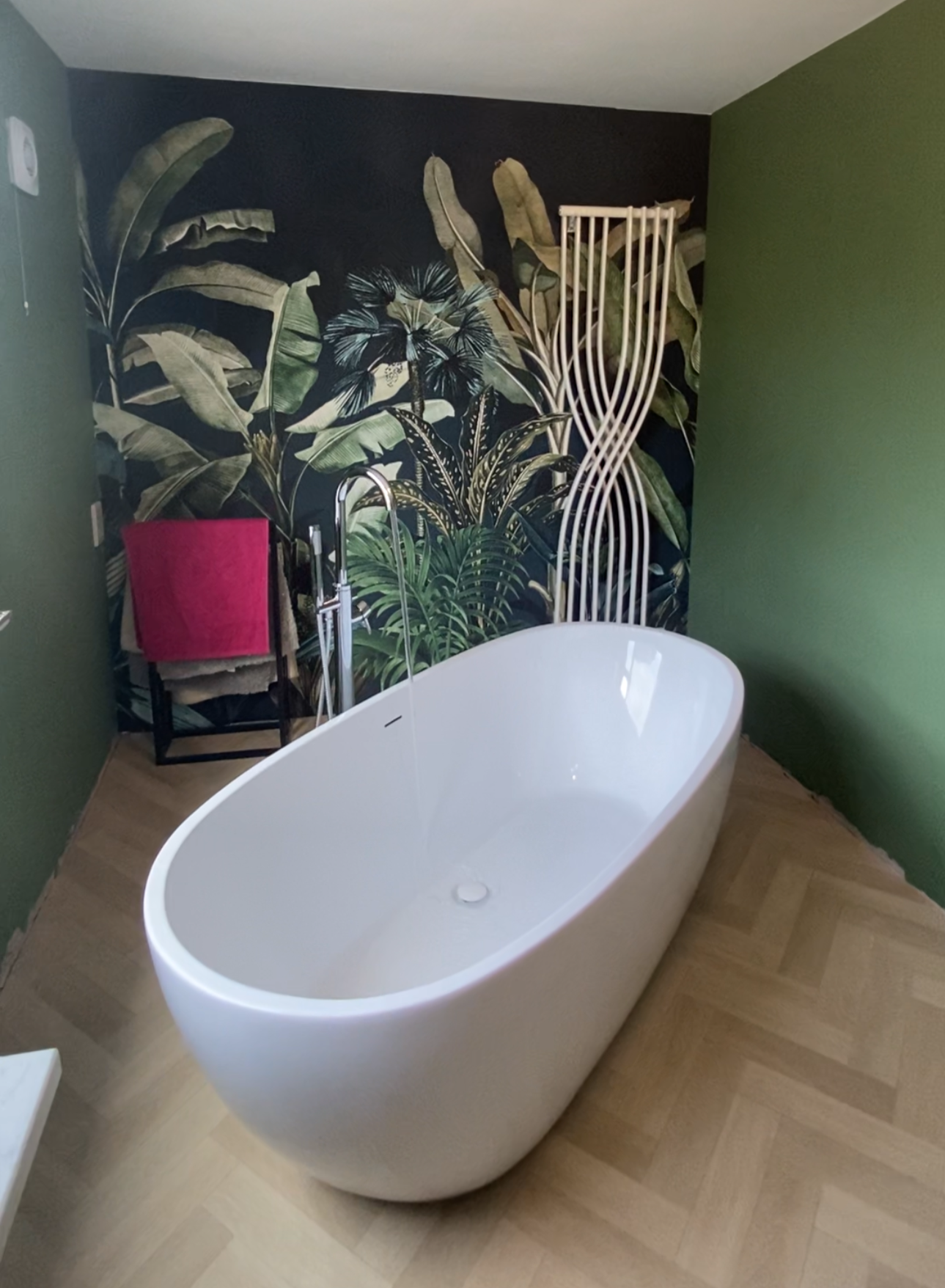 A modern bathroom with a free-standing white bathtub, tropical leaf wallpaper, a black stand with a pink towel, and green walls.