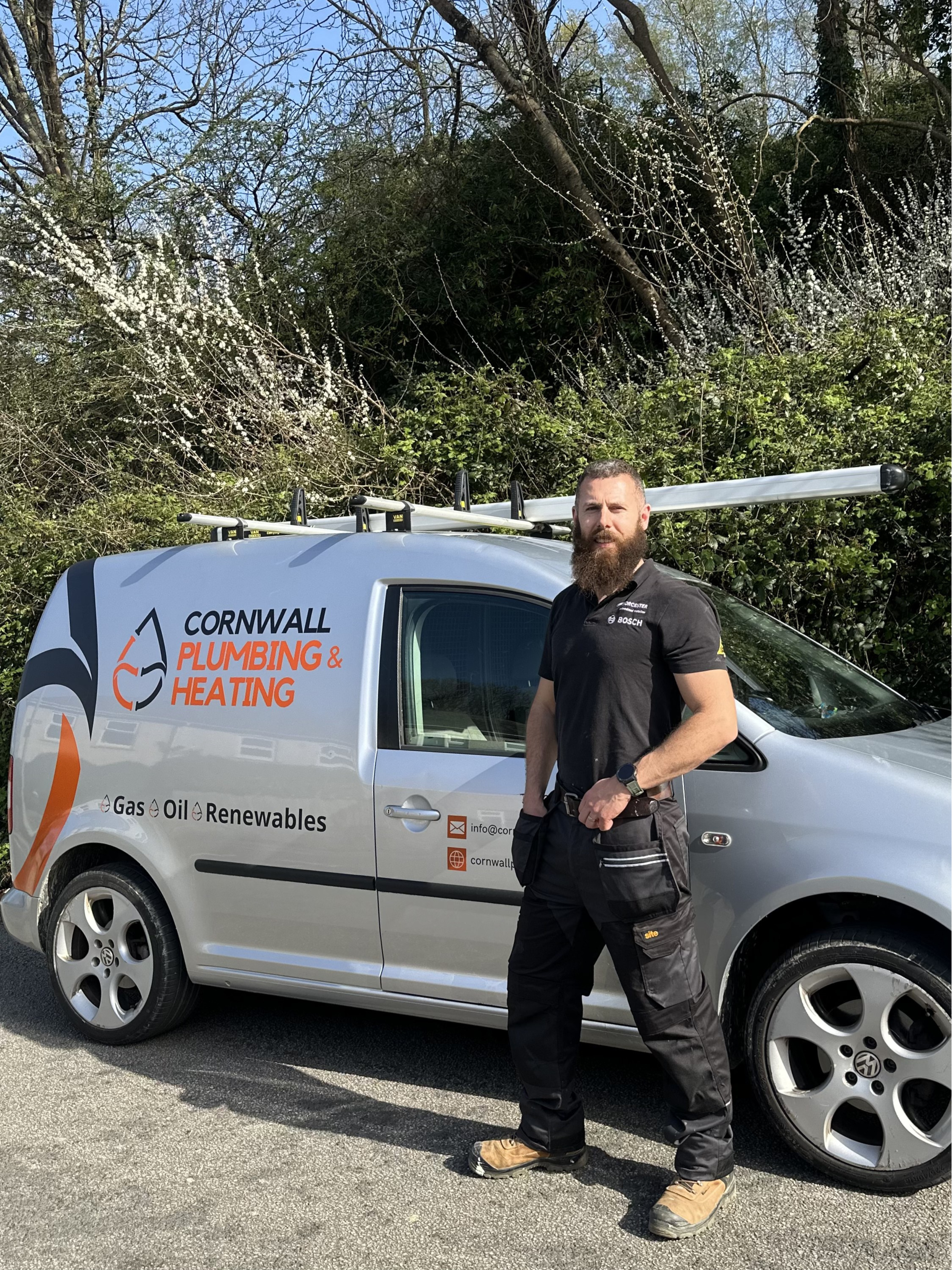 A man with a beard wearing a black uniform standing next to a gray van with 'Cornwall Plumbing & Heating' logo, parked outdoors near some trees and bushes