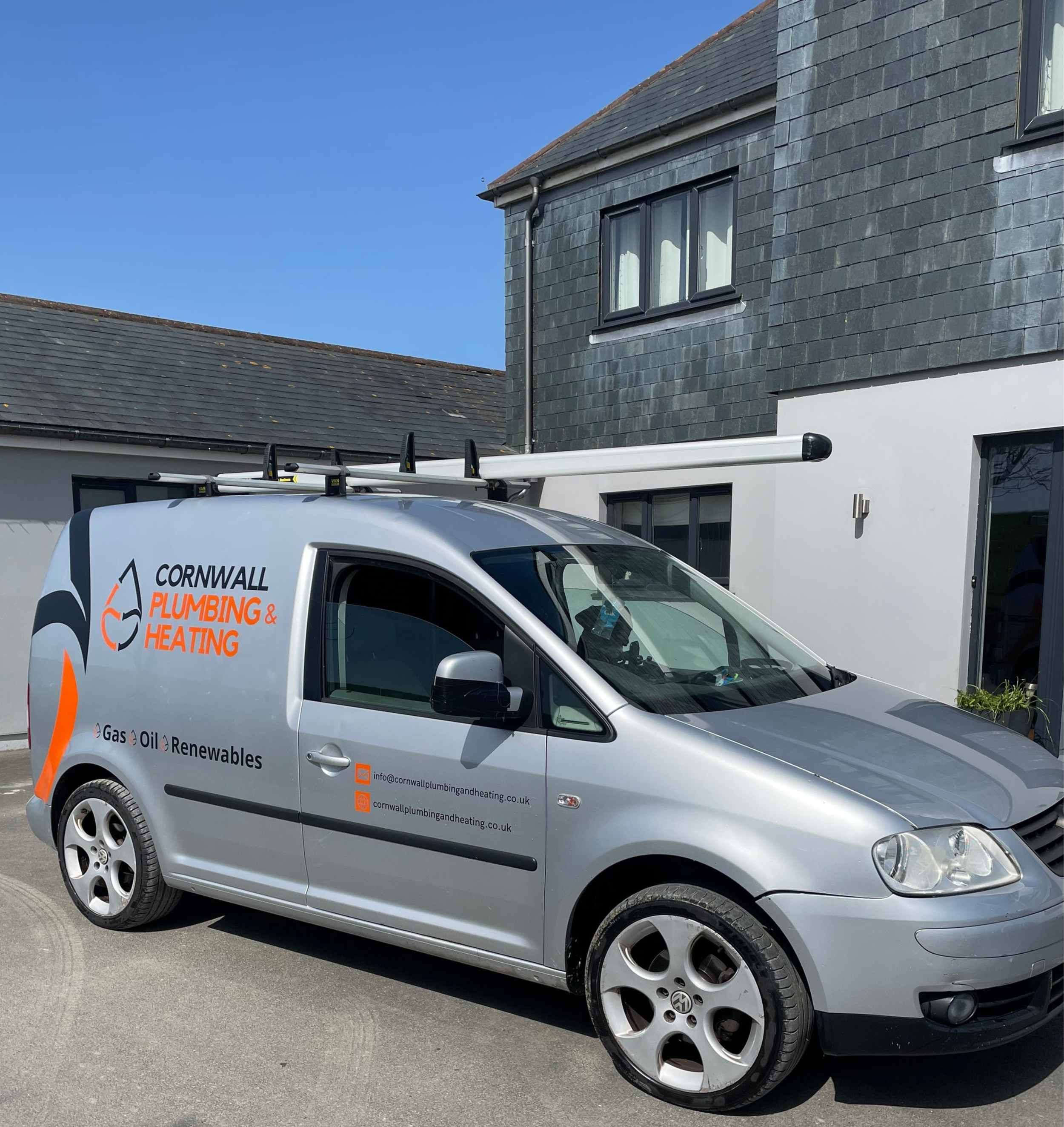 Silver service van for Cornwall Plumbing & Heating parked on a residential street under a clear blue sky, with a house featuring dark shingles and white walls in the background.