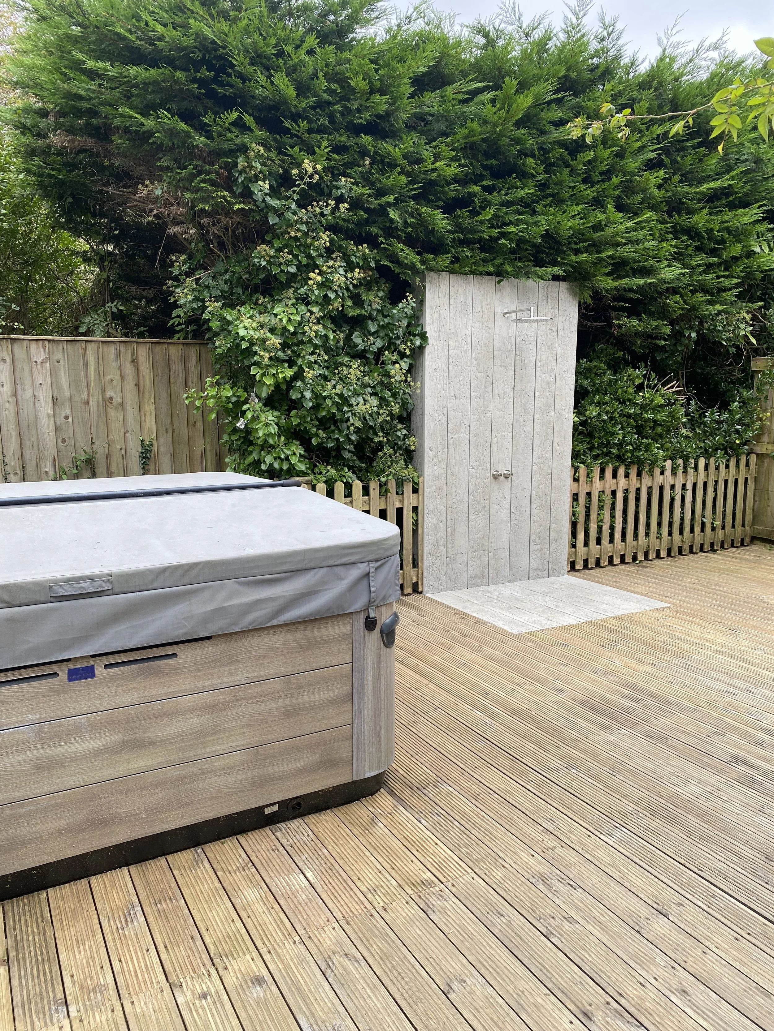 An outdoor wooden deck featuring a hot tub and a wooden shower with a latch, surrounded by green bushes and trees.