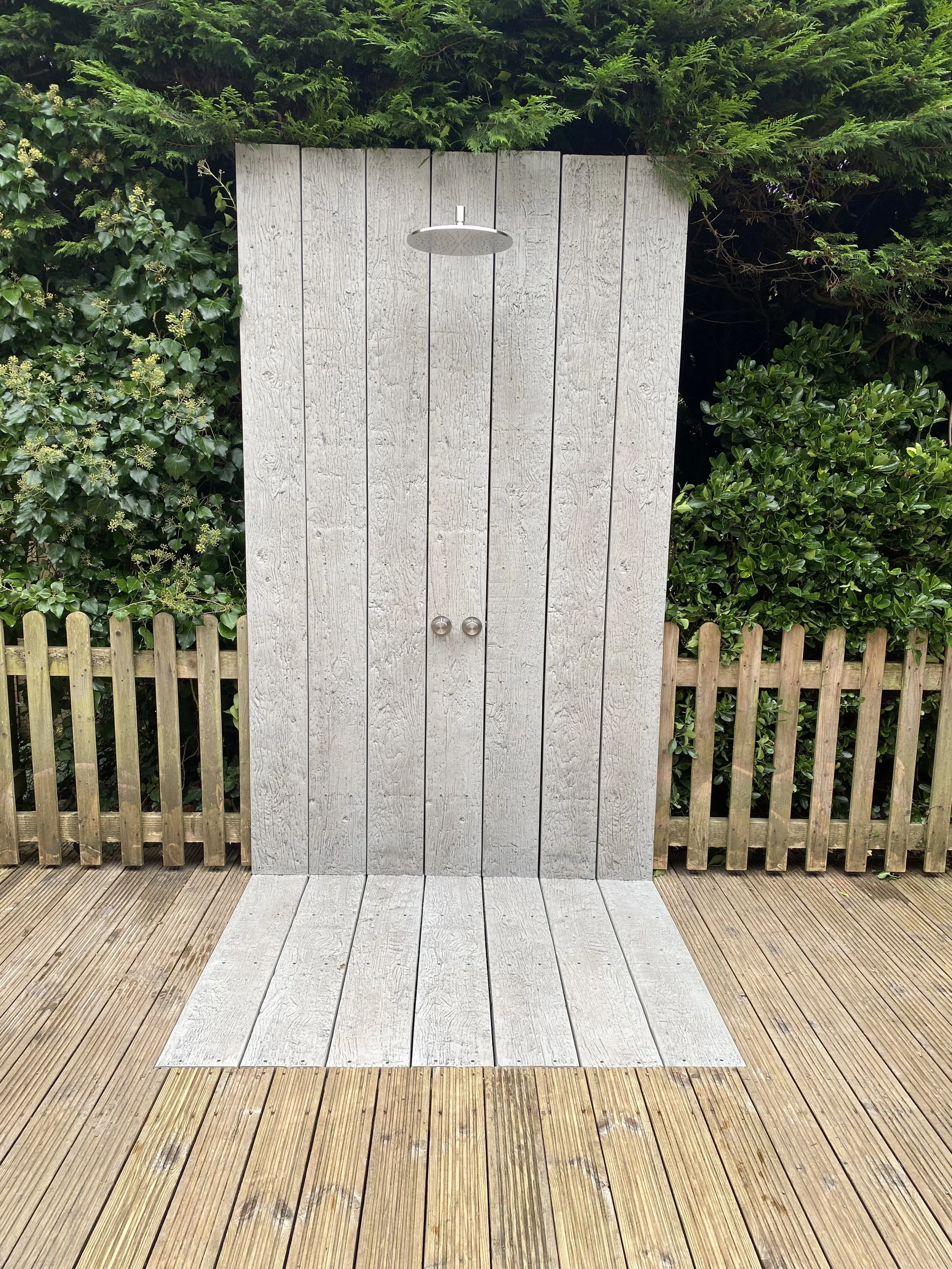 An outdoor wooden shower with a rain shower head mounted on a wooden privacy wall, surrounded by greenery and on a deck with wooden planks.