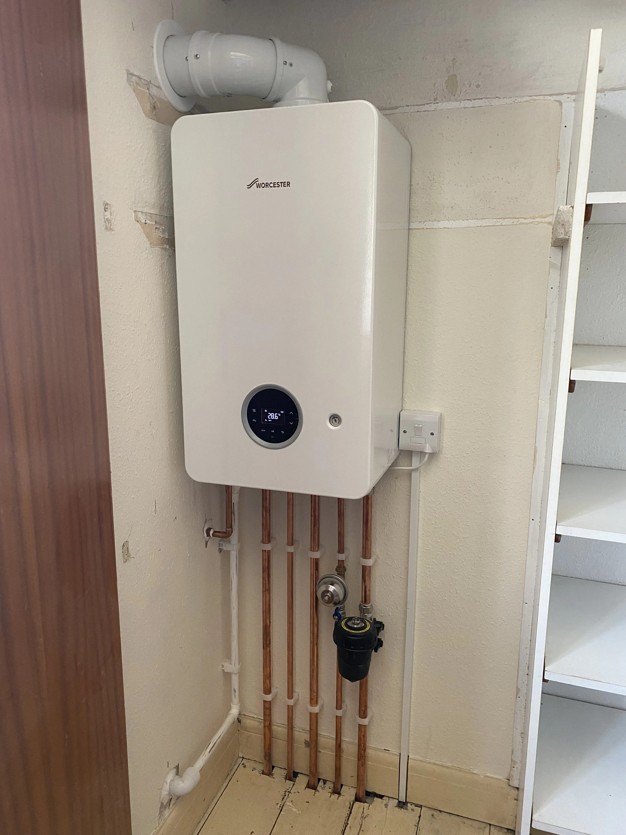 White Worcester boiler mounted on a wall in a utility room with copper pipes, a black pressure gauge, and electrical wiring.