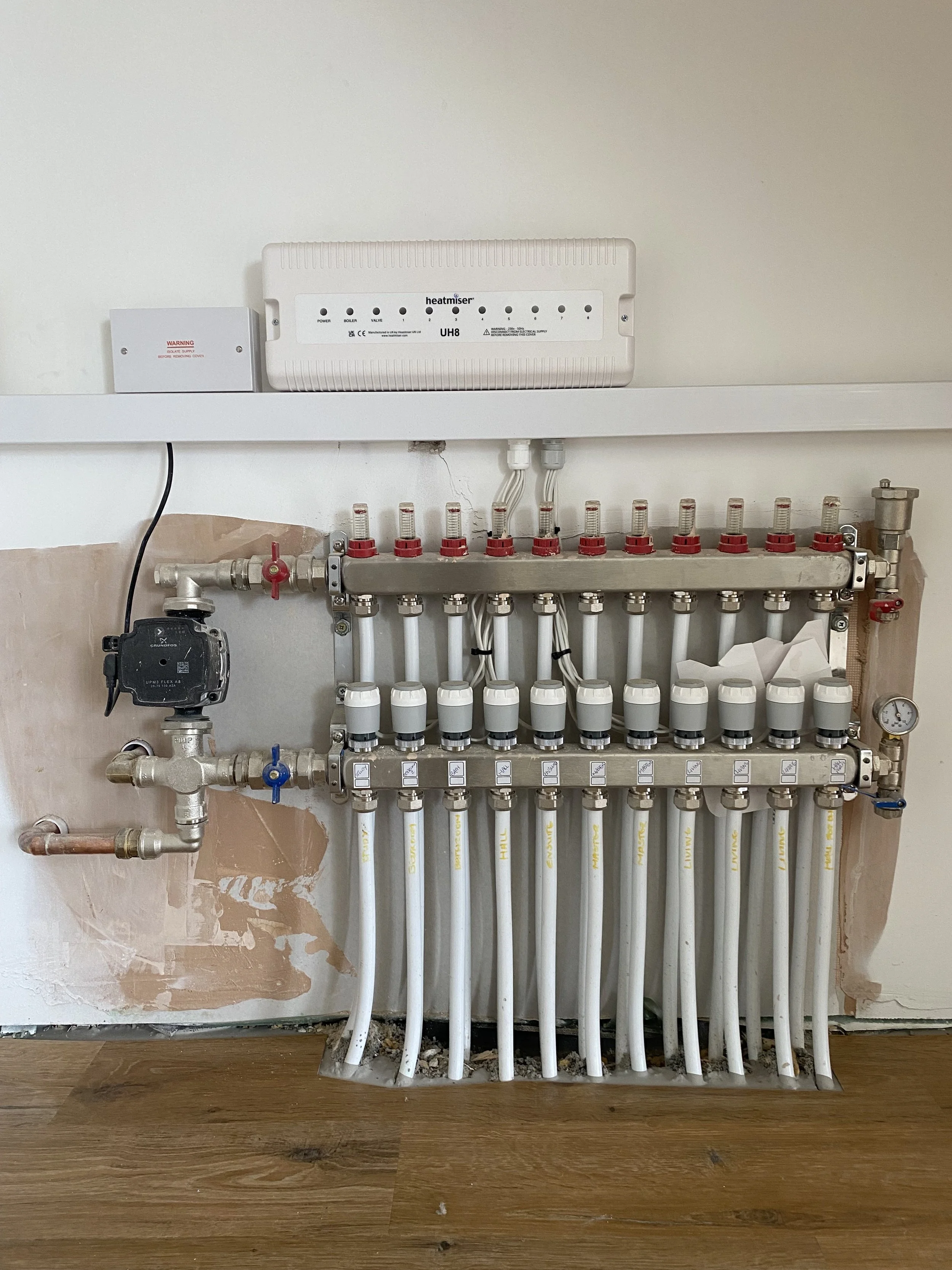 A plumbing manifold with multiple white pipes, valves, and a pressure gauge on a wall. Above it is a white digital temperature controller and a white box with a warning label.