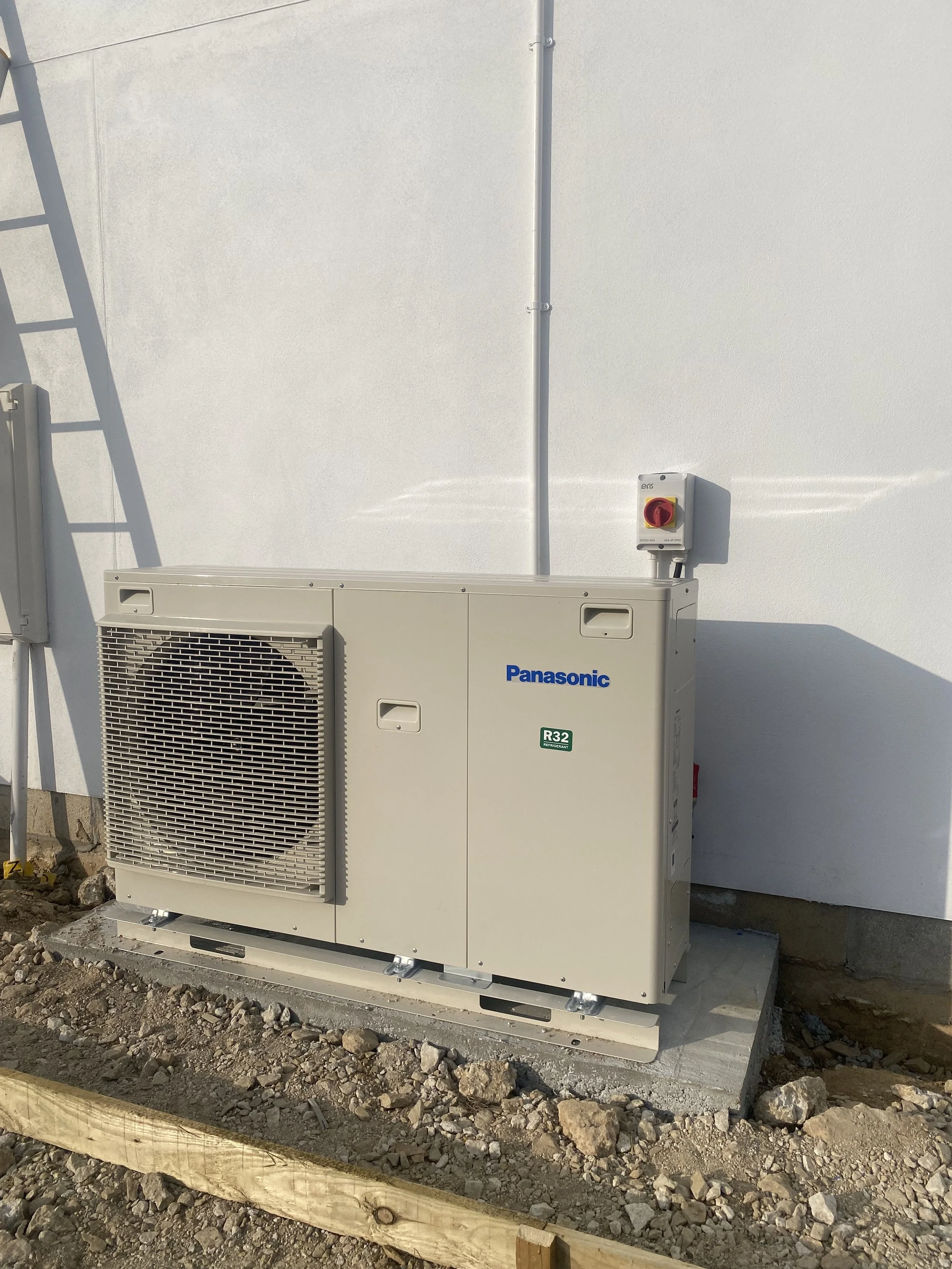 Outdoor Panasonic HVAC unit on concrete pad attached to a white wall with an electrical switch box above it. Gravel and dirt ground surrounding the unit.