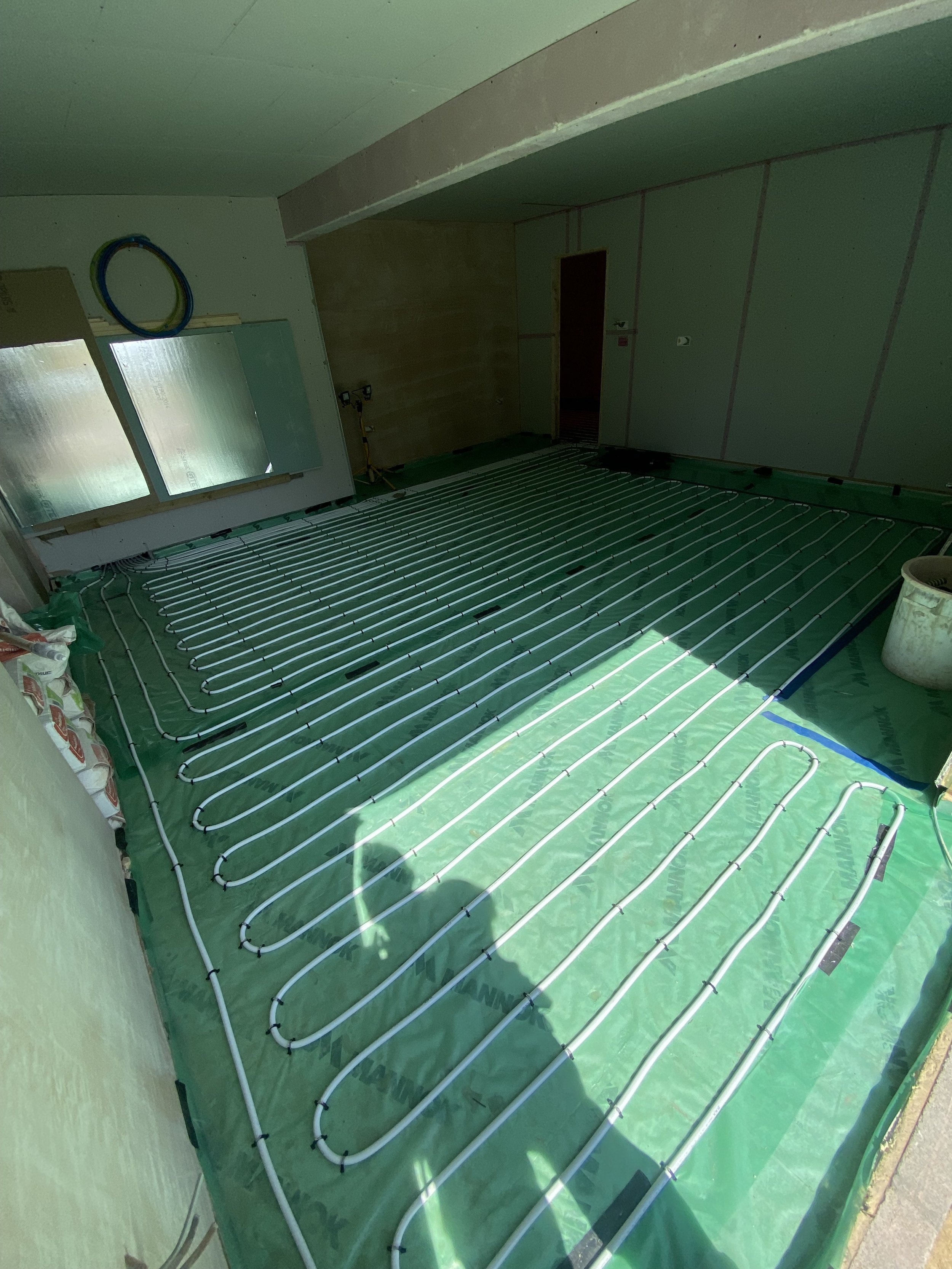 Installation of underfloor heating pipes in a room under construction, with green vapor barrier on the floor and walls.