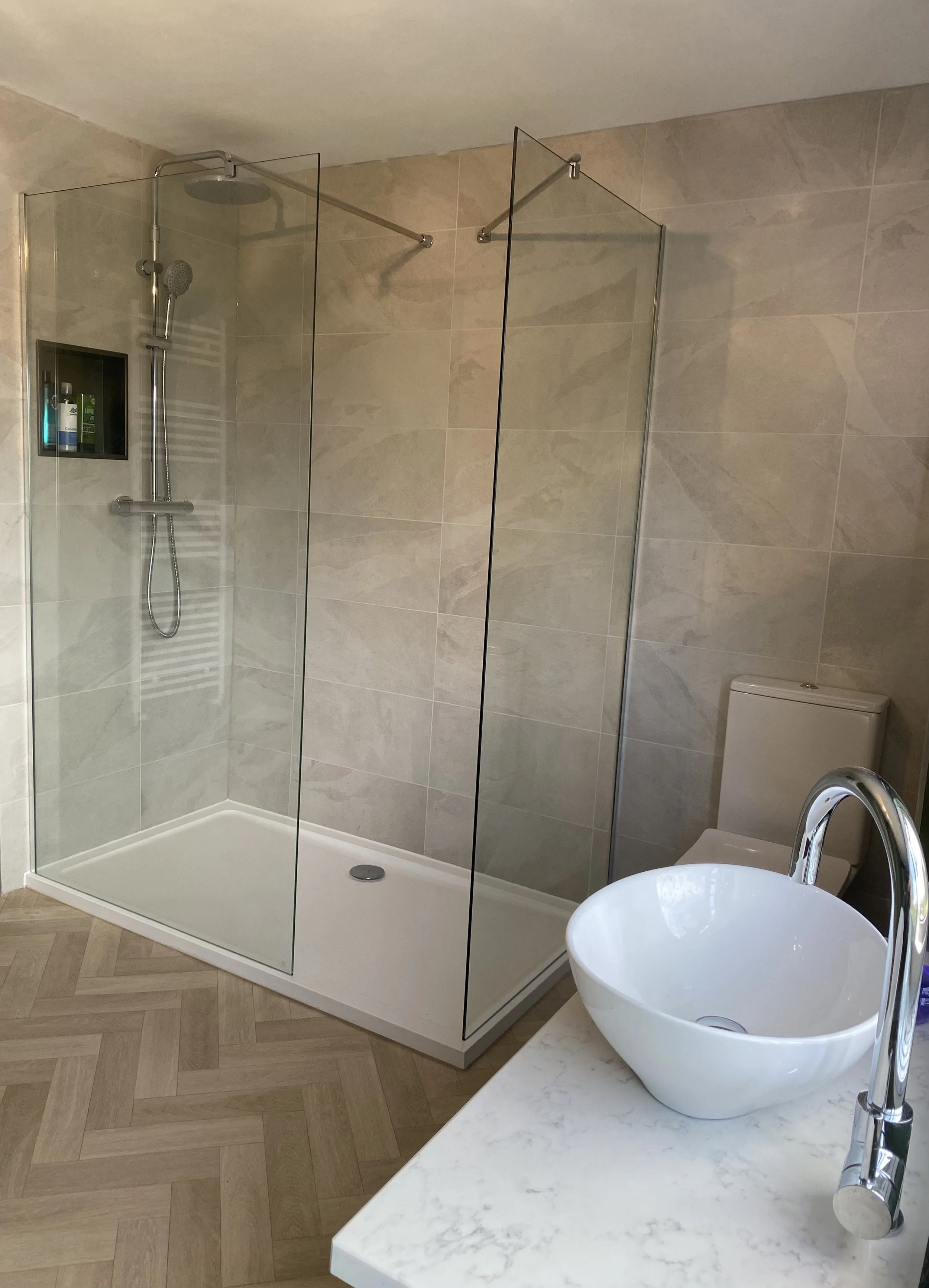 Modern bathroom with a glass-enclosed shower, a white vessel sink on a marble countertop, and a toilet, with beige tiled walls and wood-look flooring.