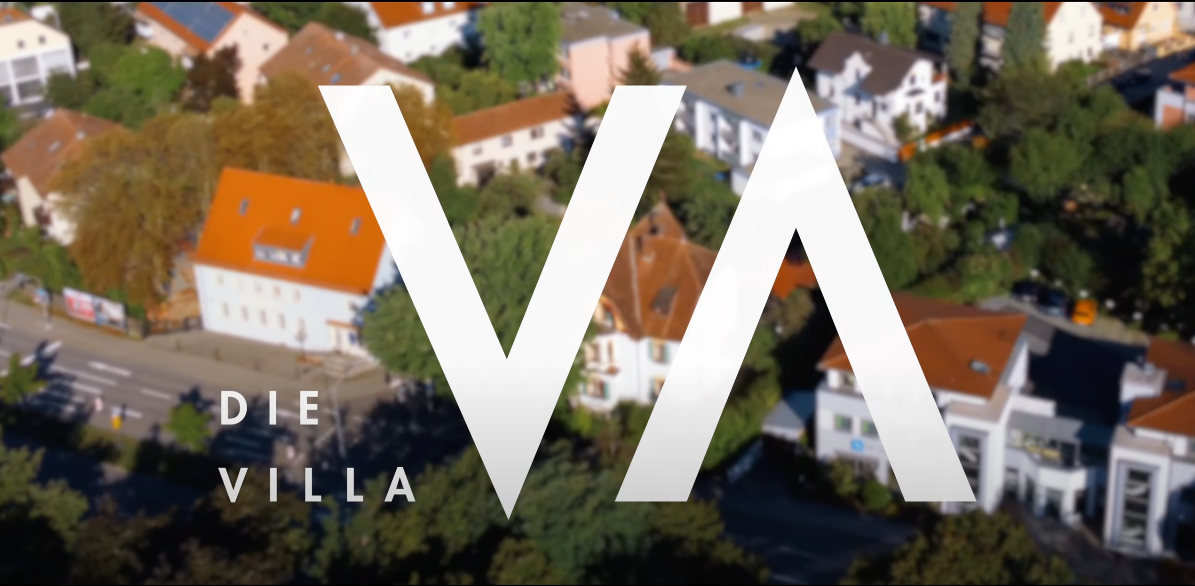 Luftaufnahme eines Wohngebietes mit zahlreichen Häusern, viel Grün und Straßen, überlagert mit einem großen weißen Logo und dem Text »Die Villa«.}
