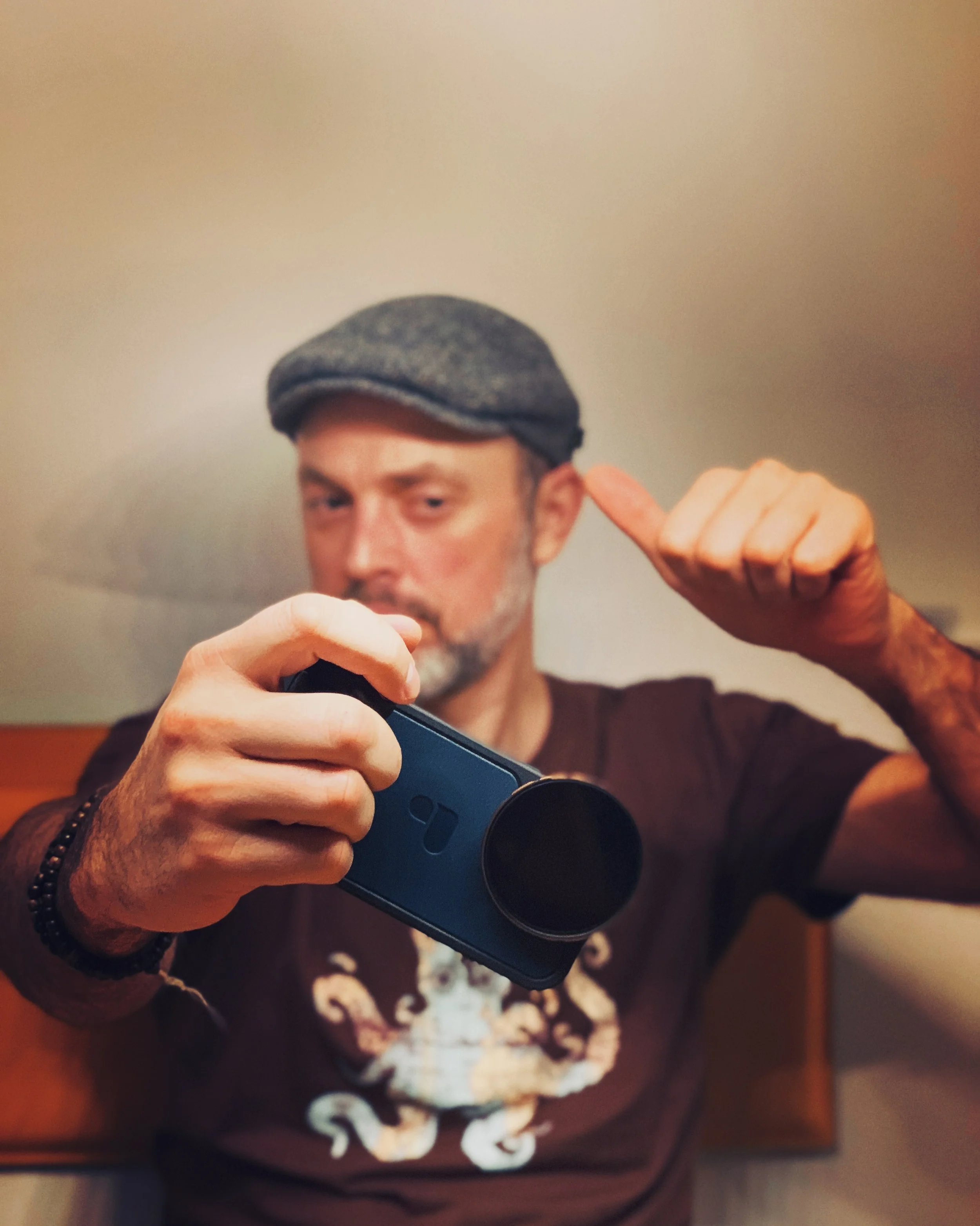 Ein Mann trägt einen grauen Hut und hält eine Kamera, während er in die Kamera fotografiert und mit der Hand auf den Kopf zeigt.