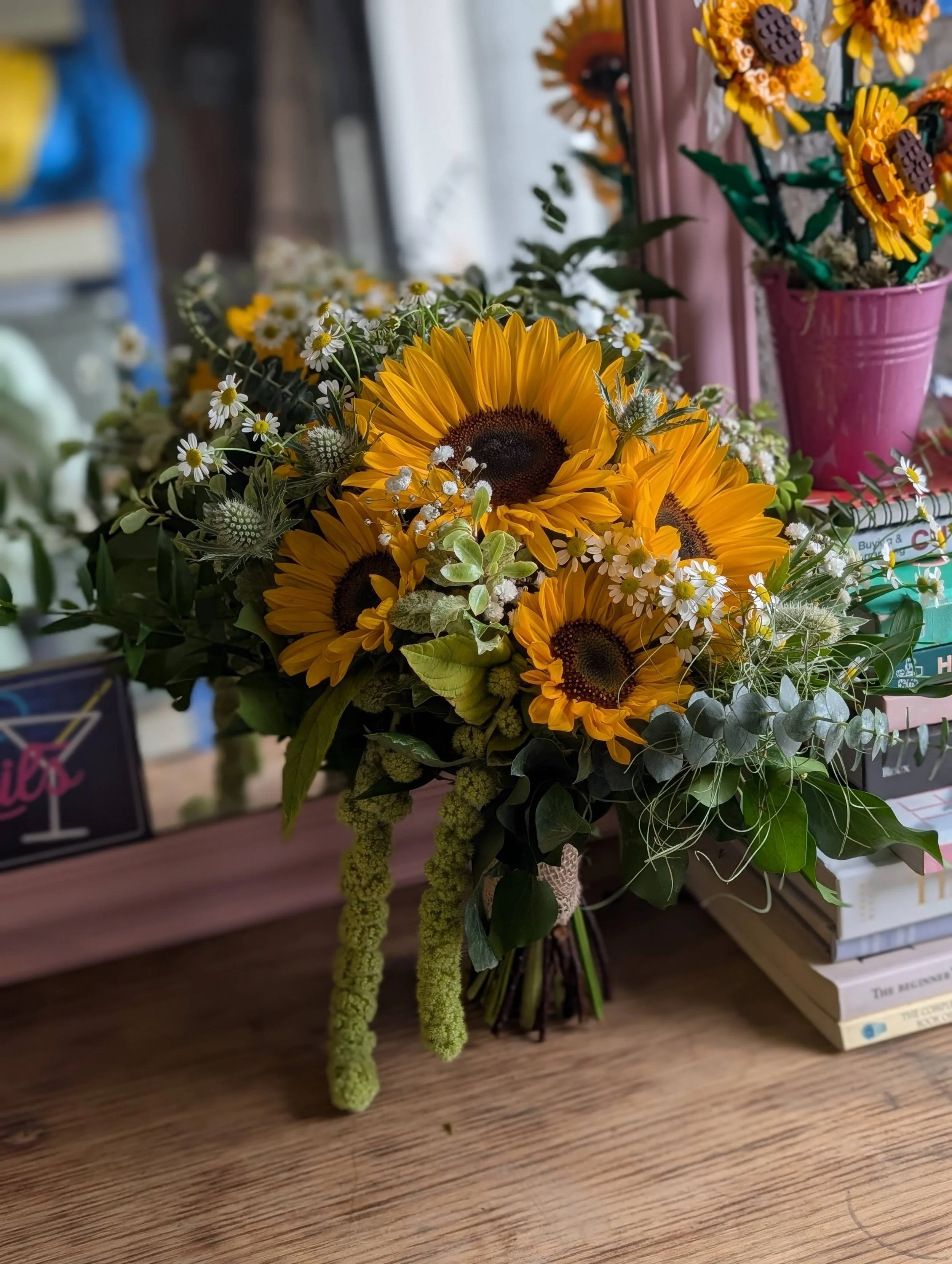 A bouquet of sunflowers and small white daisies on a wooden surface, with other floral arrangements and books in the background.