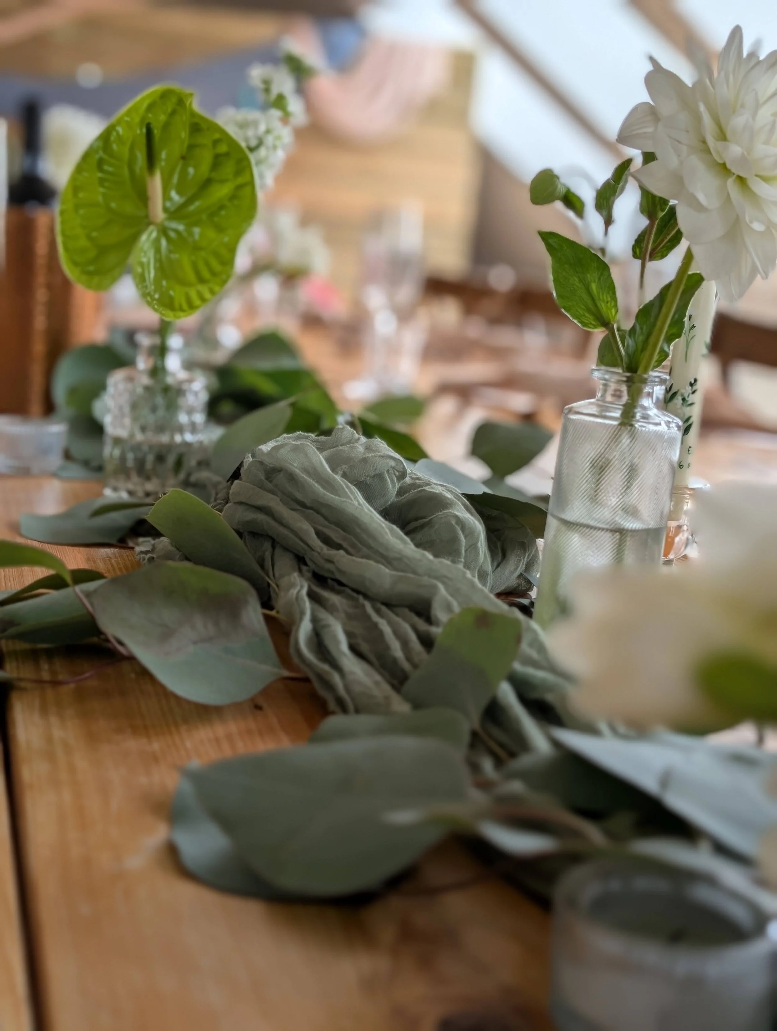 Decorative table setting with a wooden table, glass vases with white and green flowers, eucalyptus leaves, and linens, in a well-lit indoor space.