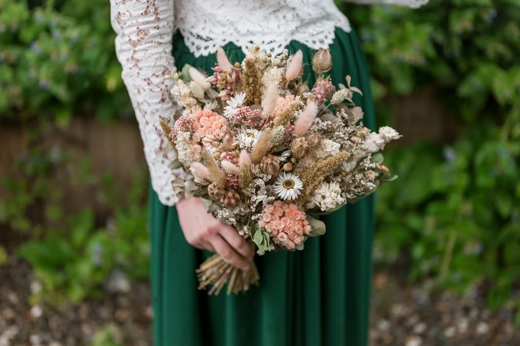 Person holding a large dried flower bouquet outdoors, with green foliage in the background, wearing a white lace top and green skirt.