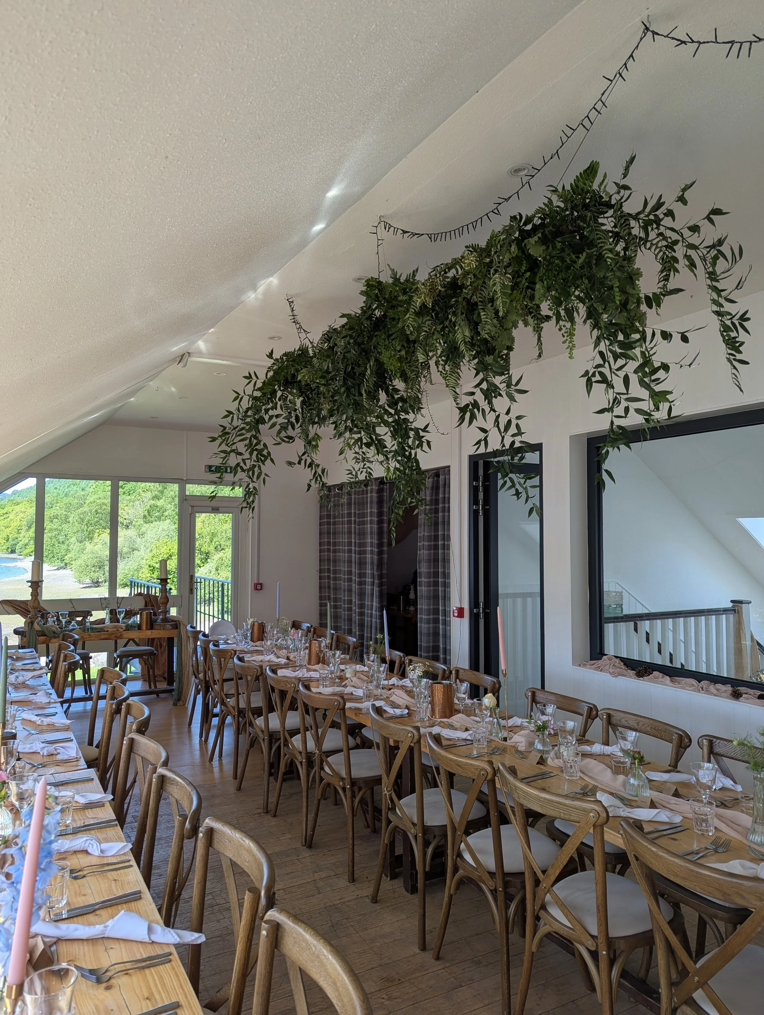 Indoor dining area set up for a gathering, with long tables covered in beige tablecloths, napkins, glassware, and small vases, decorated with greenery, with large windows showing a view of trees and water outside, and hanging greenery from the ceilin
