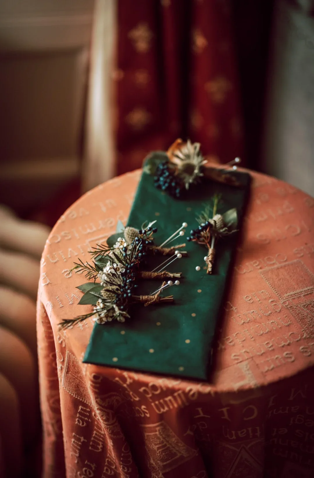 Decorative boutonniere with pine branches, berries, and white flowers on a green fabric, placed on a pink tablecloth.