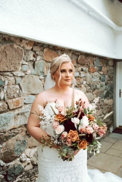 Bride in wedding dress and tiara holding a large bouquet of flowers, standing outside against a stone wall and white building.
