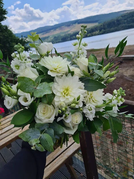 A bouquet of white flowers including roses and dahlias, with green leaves, held outdoors near a lake with hills in the background.