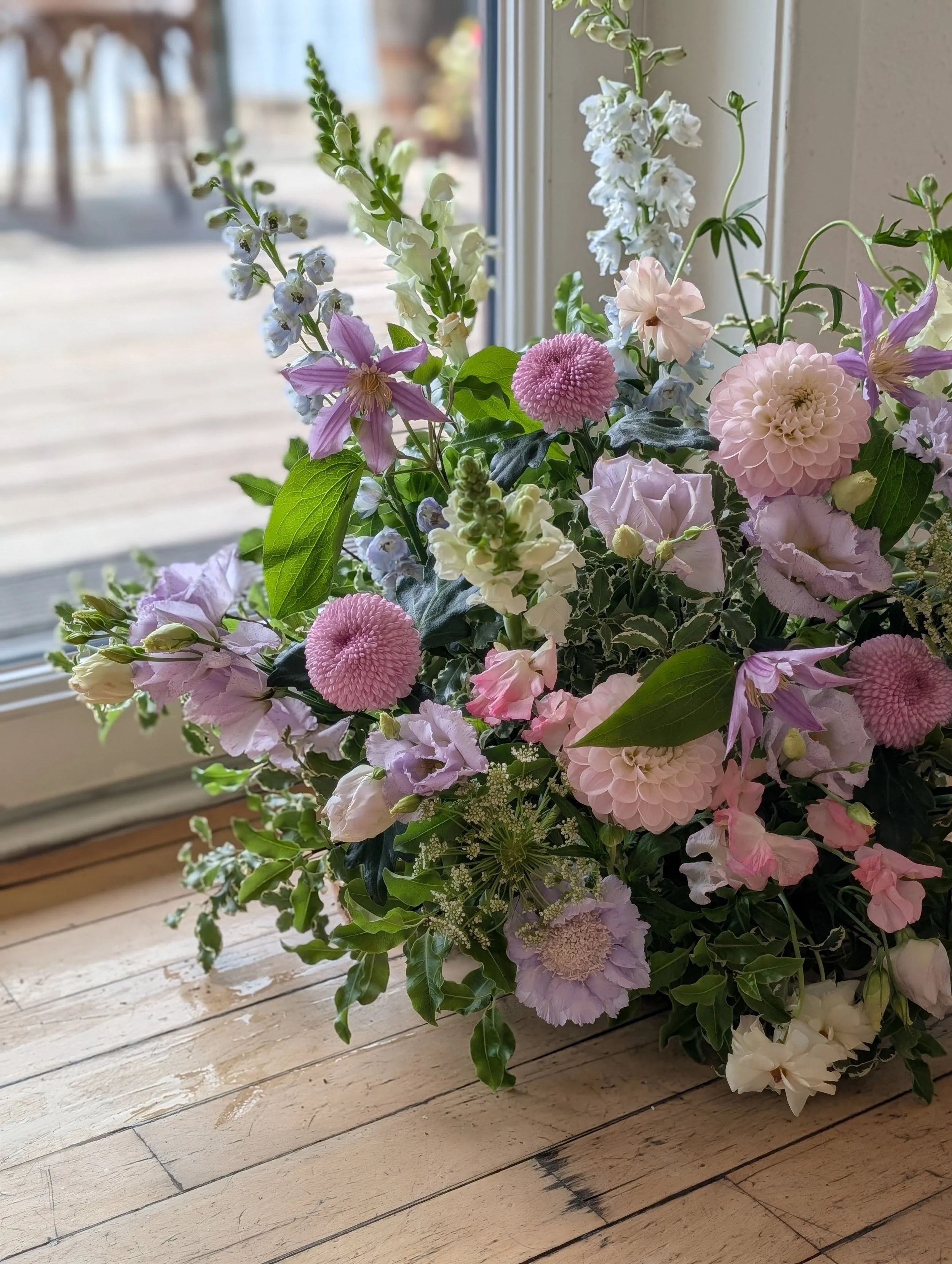 A bouquet of pastel pink, purple, and white flowers, placed on a wooden surface near a window, with a blurred outdoor scene in the background.