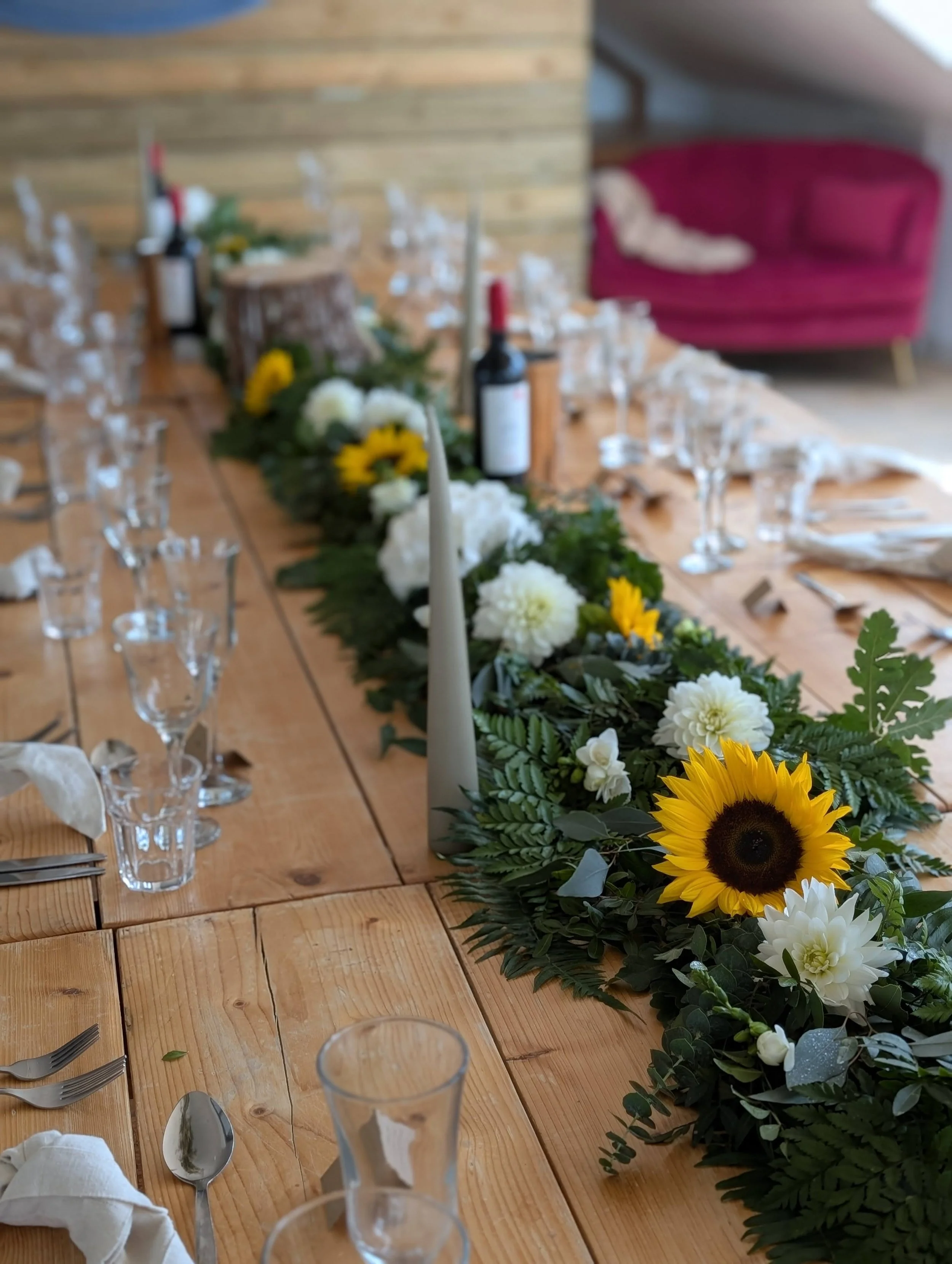 A long wooden dining table decorated with a floral centerpiece featuring sunflowers, white and yellow flowers, and green foliage, set with wine glasses, silverware, and white napkins, in a room with wooden wall panels, a pink velvet sofa in the backg