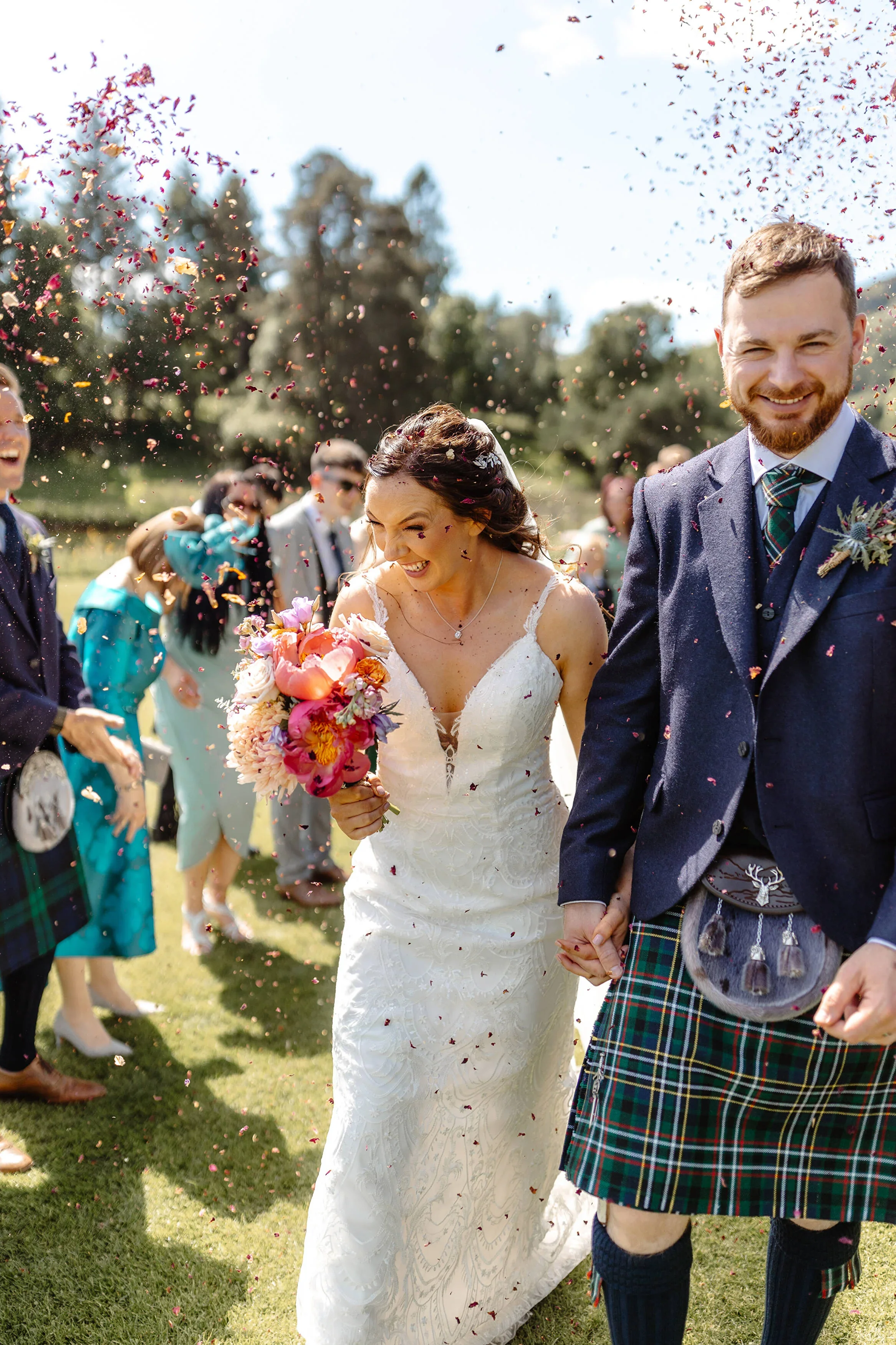 A bride and groom walking hand-in-hand through confetti with guests celebrating around them outdoors on a sunny day.