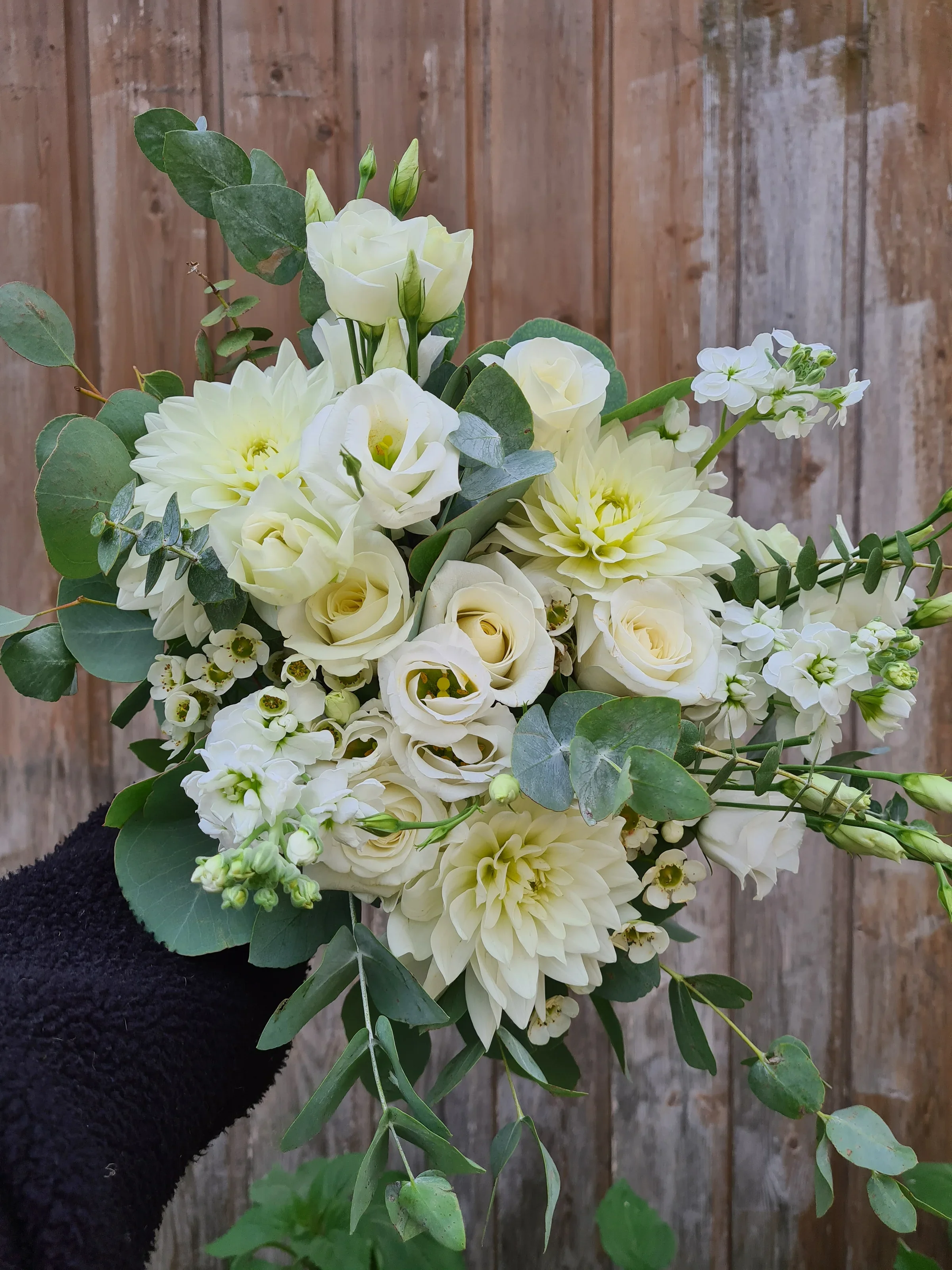 A bouquet of white flowers including roses, dahlias, and smaller blossoms with green leaves, held in front of a wooden fence.