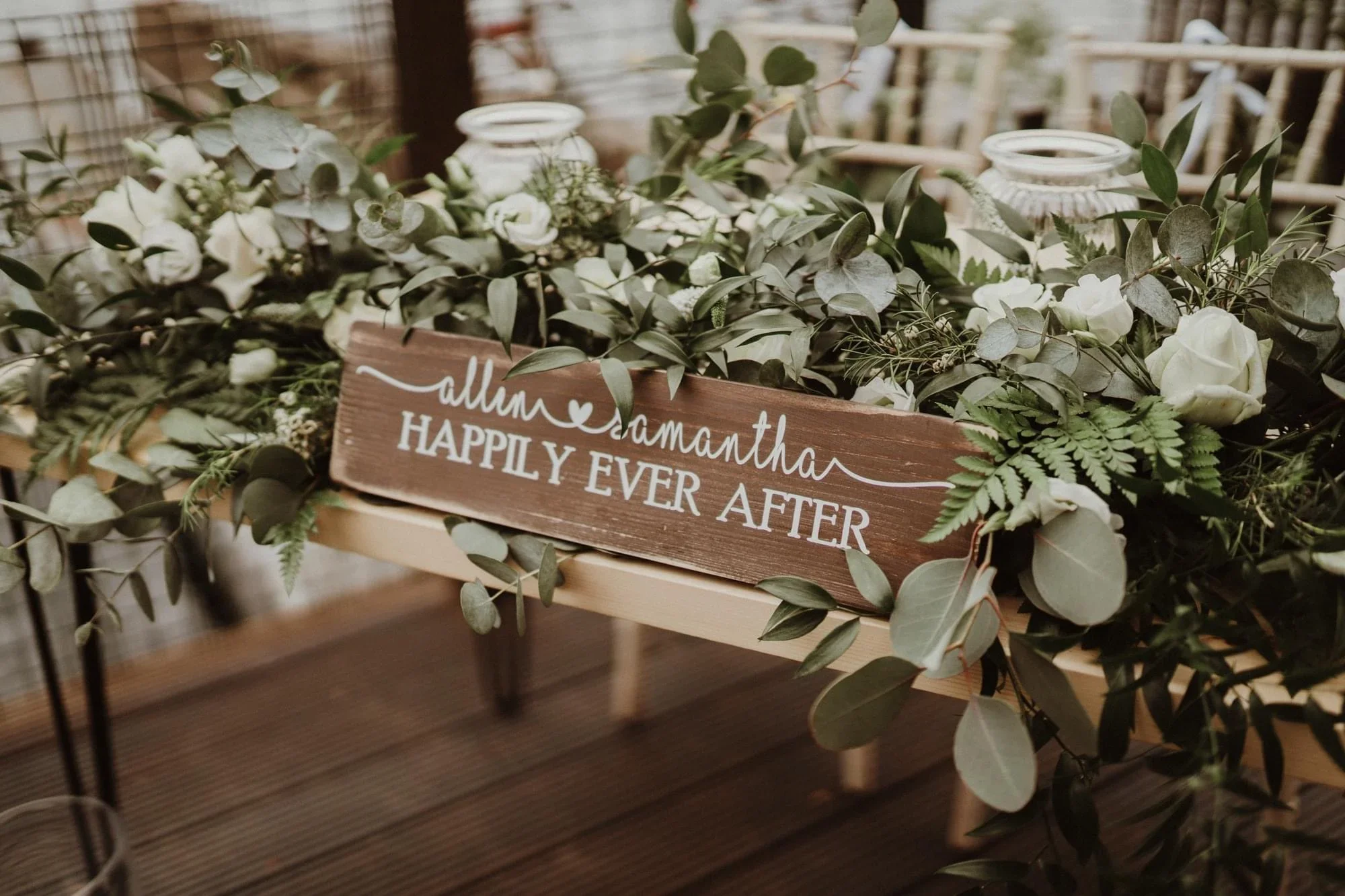 A wooden sign placed in a floral arrangement with white roses and greenery, reading 'Allan & Samantha Happily Ever After' at a wedding or celebration event.
